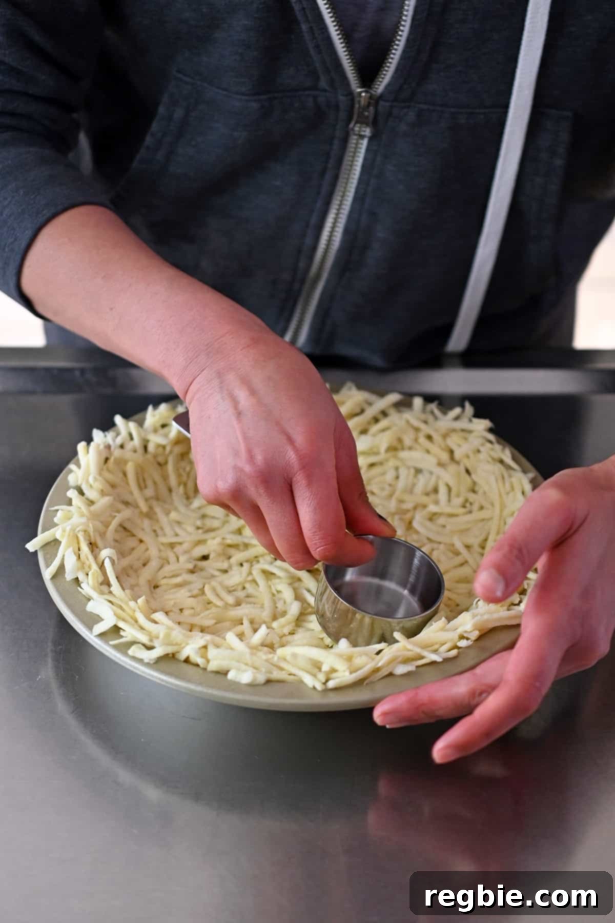 A hand using a measuring cup to firmly press shredded potatoes into an even, consistent layer within a pie plate, forming the quiche crust.