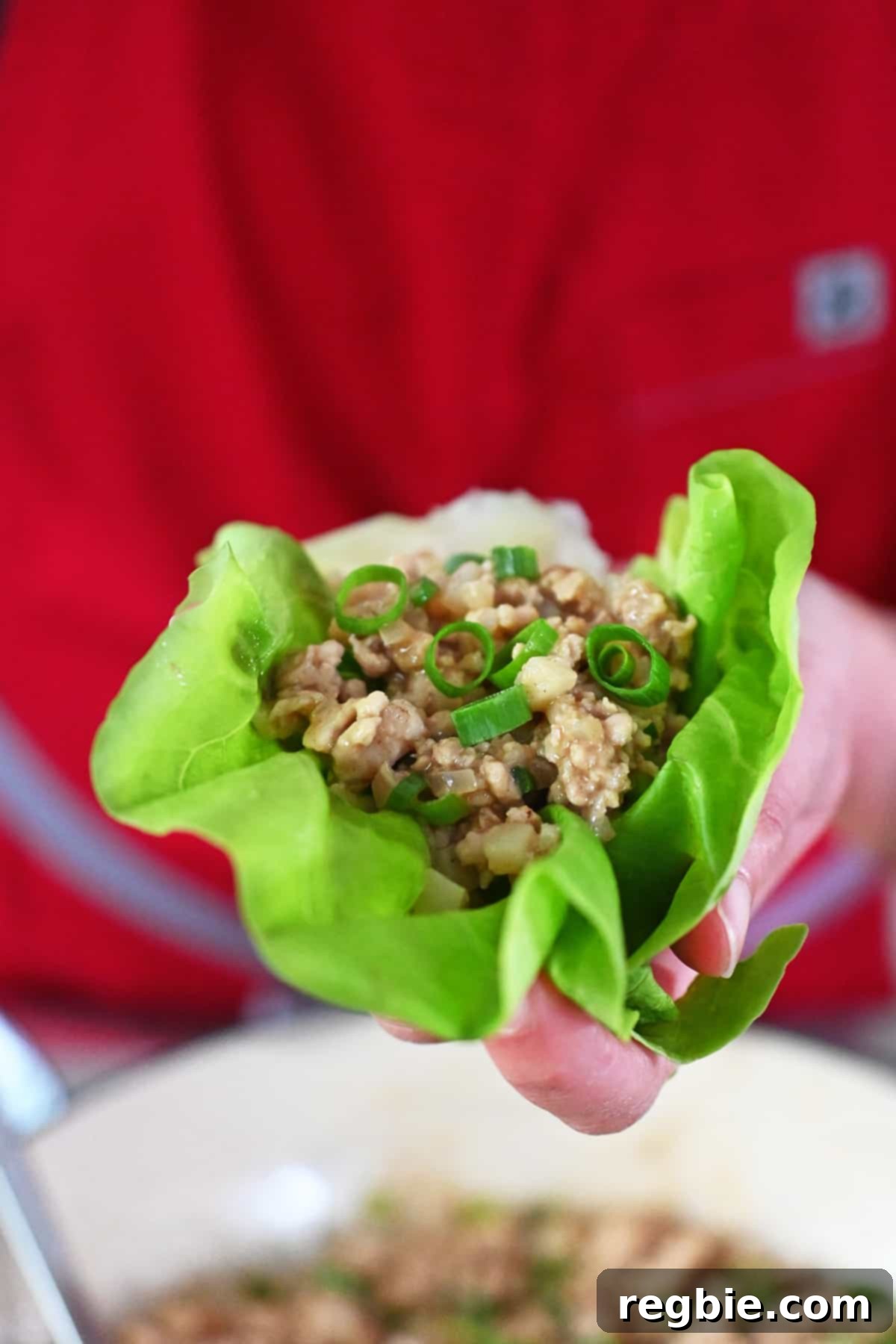 A hand holding a chicken lettuce wrap over a skillet filled with the savory filling.