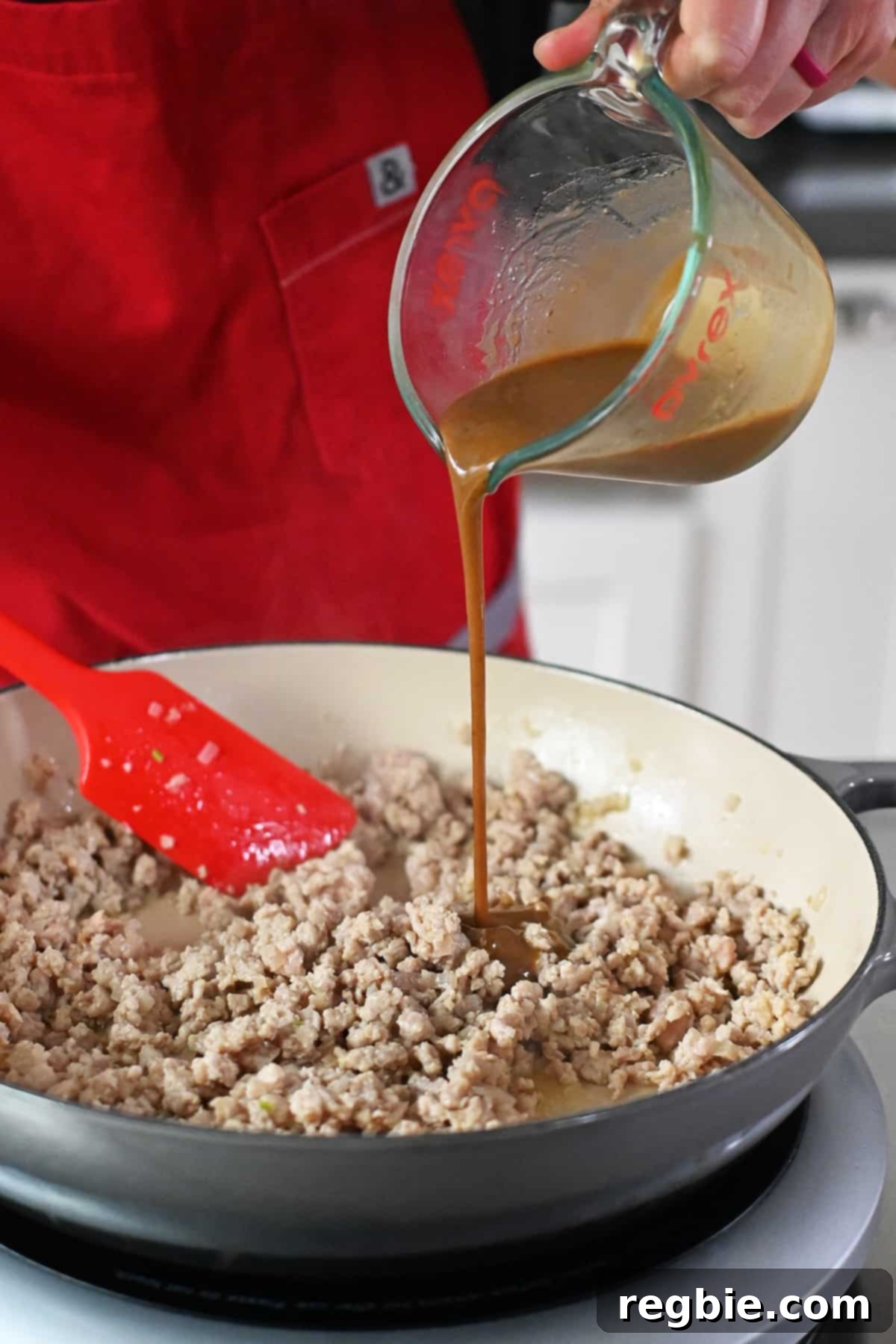 Pouring a brown sauce from a measuring cup into a skillet with cooked ground chicken.