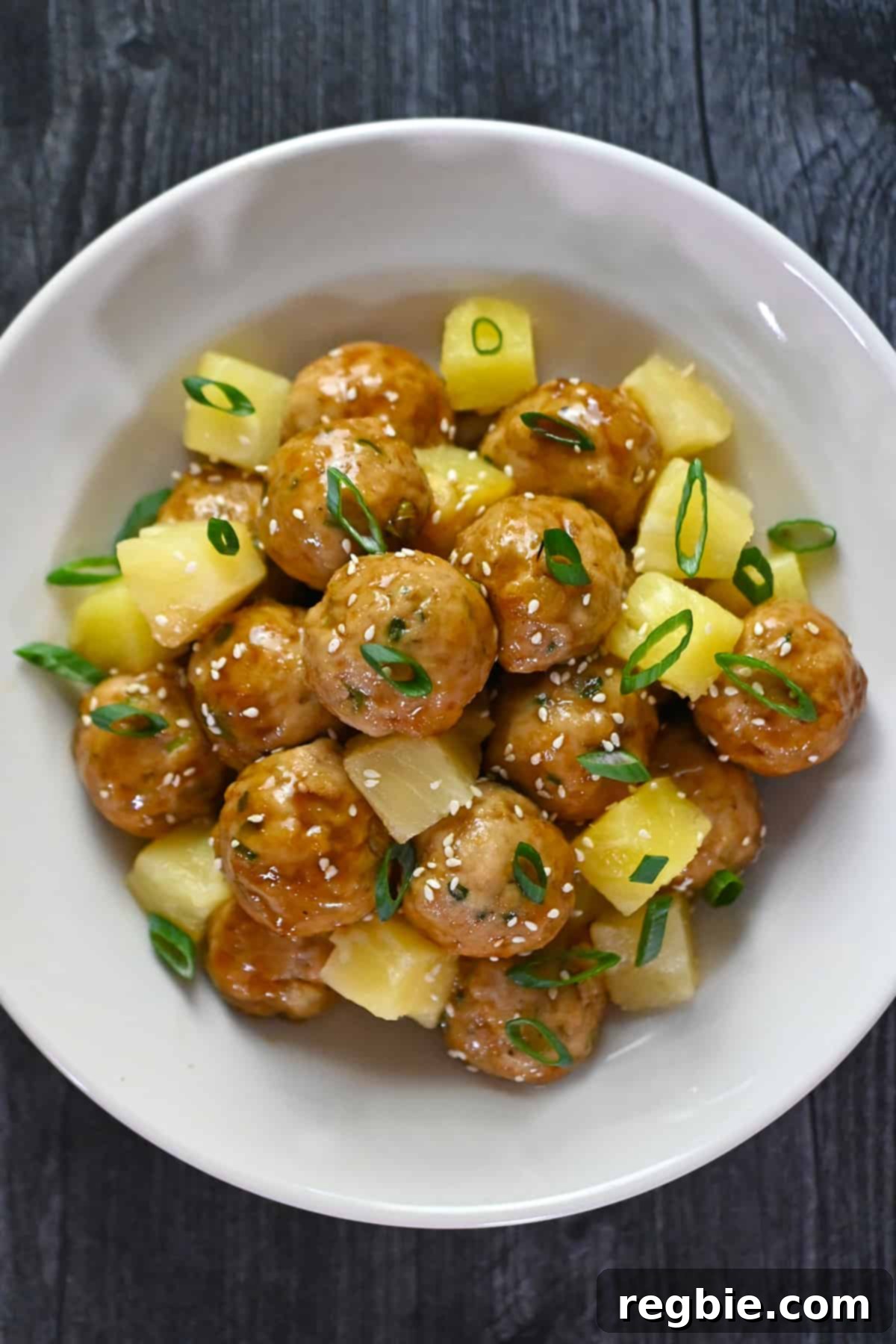 An overhead shot of a white plate filled with teriyaki pineapple meatballs and cubed pineapple, topped with toasted sesame seeds and sliced green onions.
