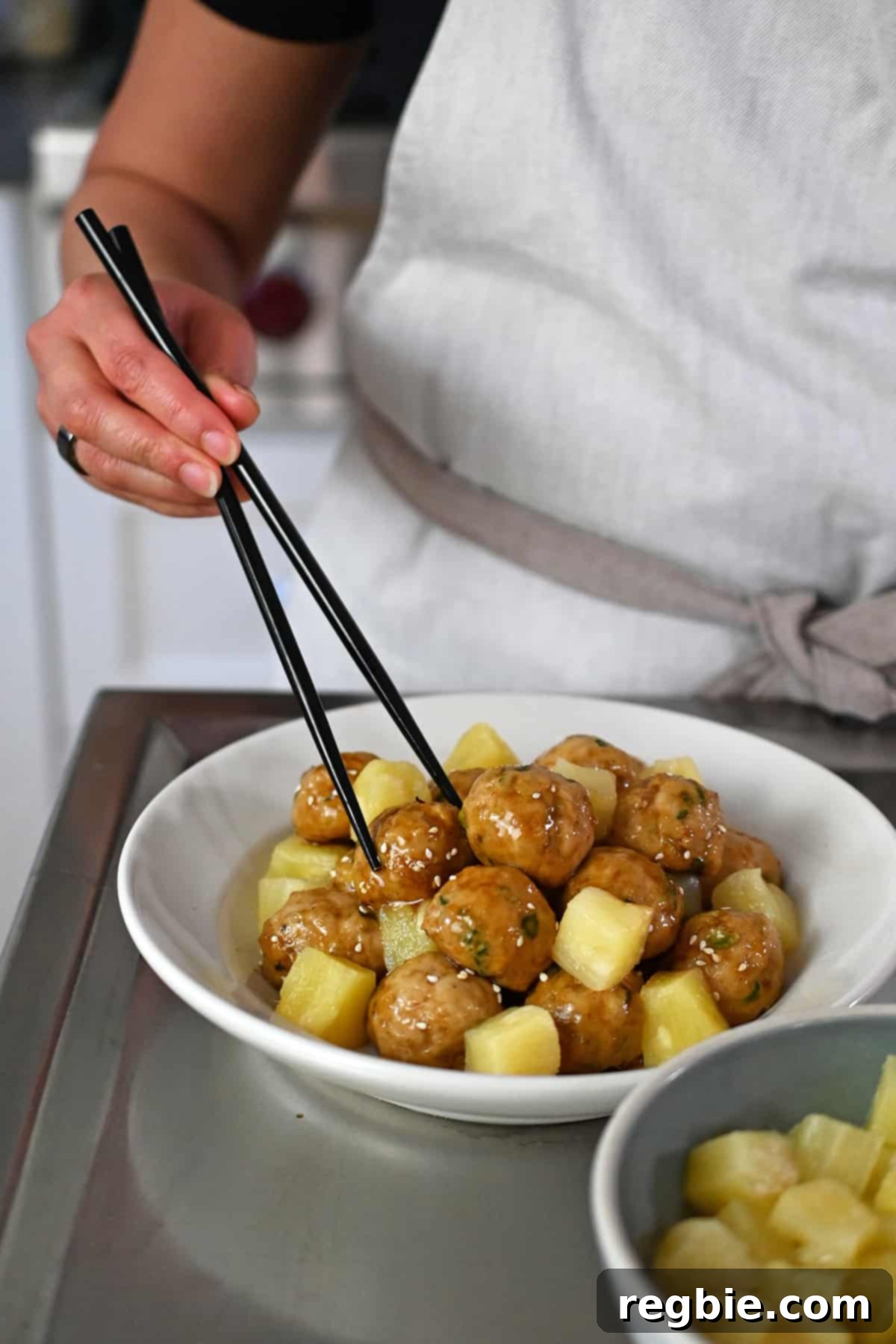A pair of chopsticks is grabbing a juicy chicken meatballs coated in sticky teriyaki sauce with pineapple chunks.