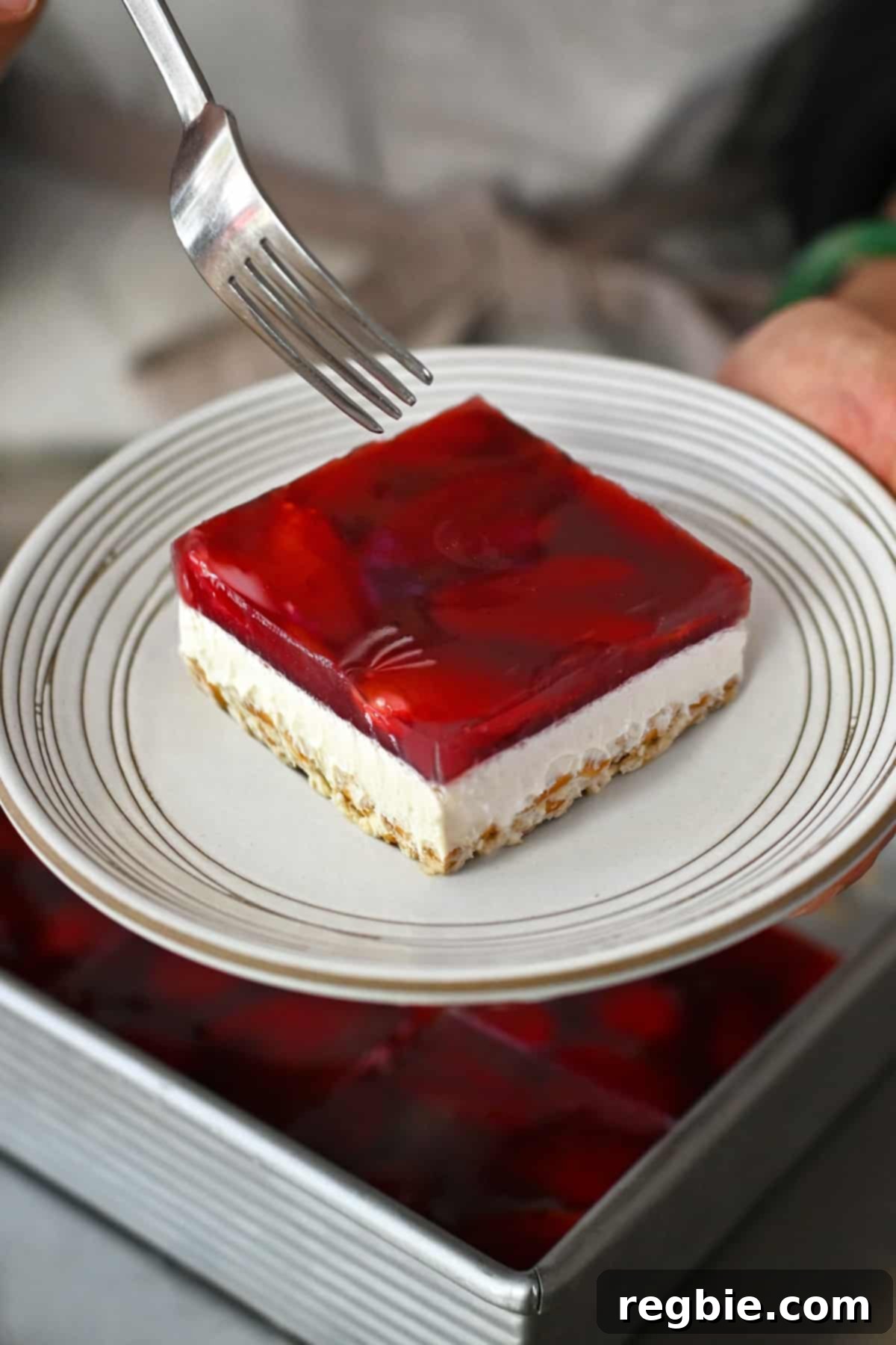A fork is hovering above a slice of gluten-free, grain-free, and dairy-free strawberry pretzel salad on a white plate, showcasing its distinct layers.