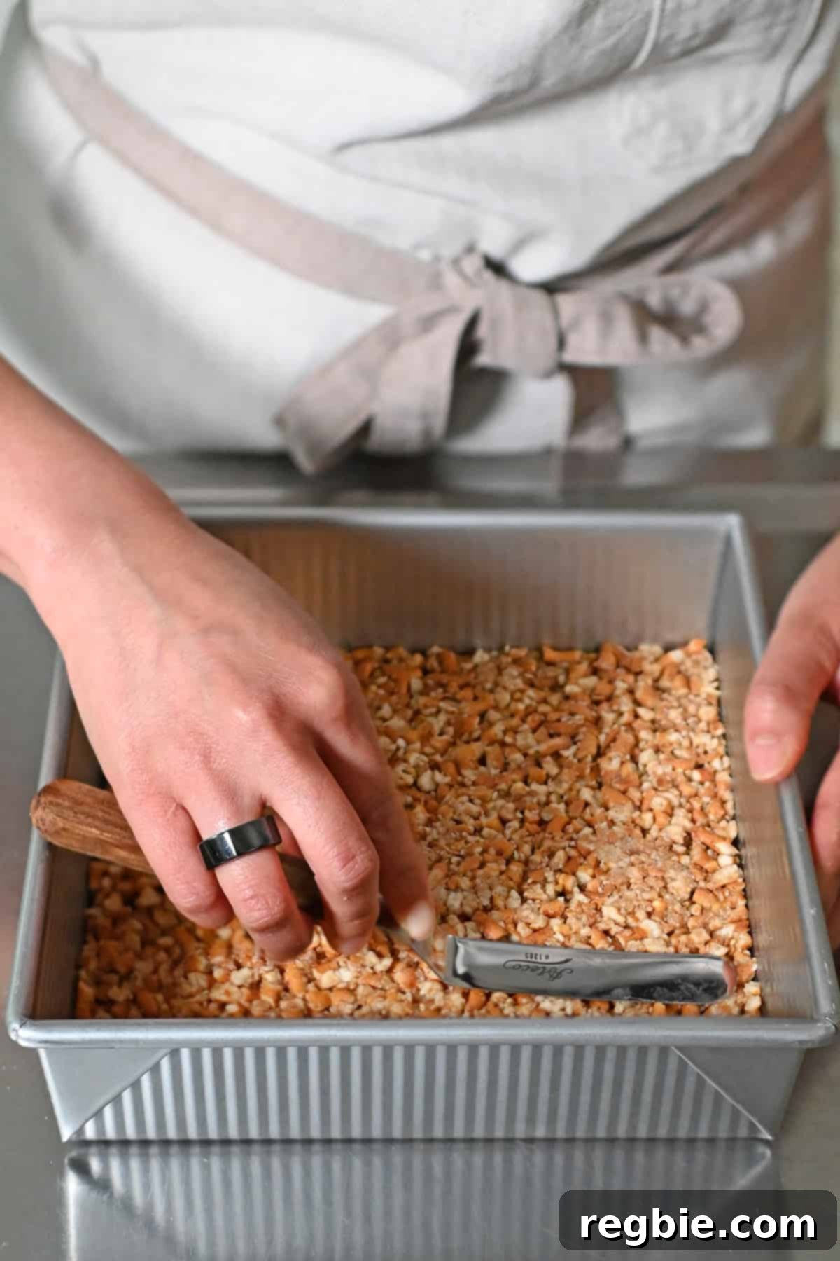 A hand uses a small offset spatula to meticulously press the pretzel crust into an even, compact layer at the bottom of a square baking pan.