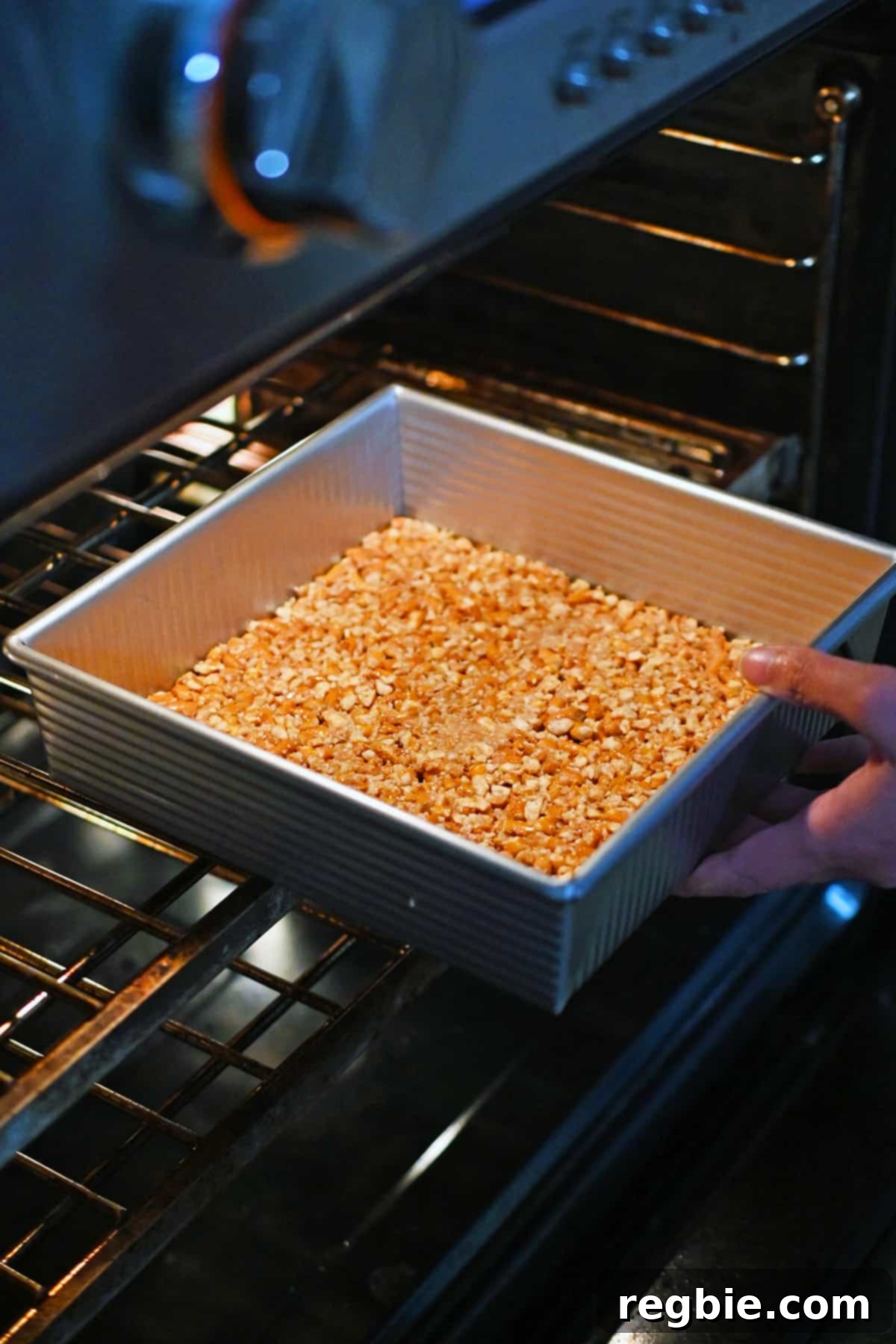 A square baking pan containing a golden-brown, baked gluten-free pretzel crust is carefully placed into an open oven, ready for the next layer.