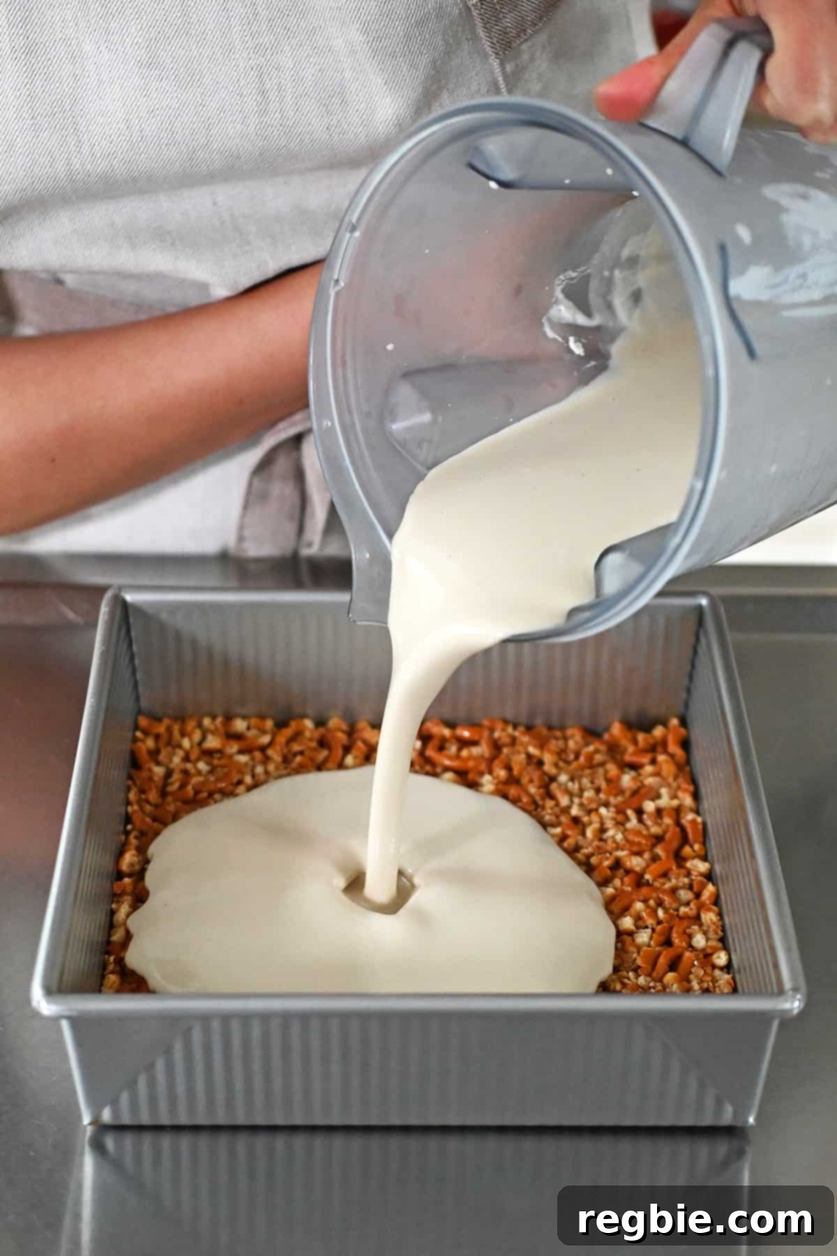 Pouring a smooth, creamy filling from a blender onto a cooled grain-free pretzel crust in a square baking pan, forming the middle layer.