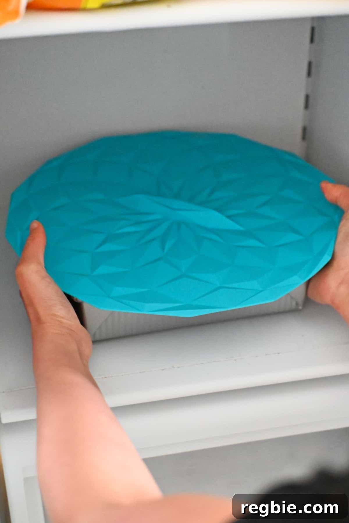 A square baking pan filled with the first two layers of strawberry pretzel salad is covered with a blue silicone lid and placed into a freezer to solidify.