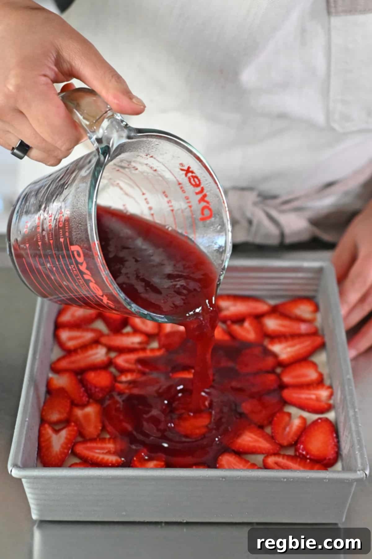 Pouring homemade strawberry gelatin from a liquid measuring cup evenly over the strawberries and creamy layer of a strawberry pretzel salad in a square baking pan.