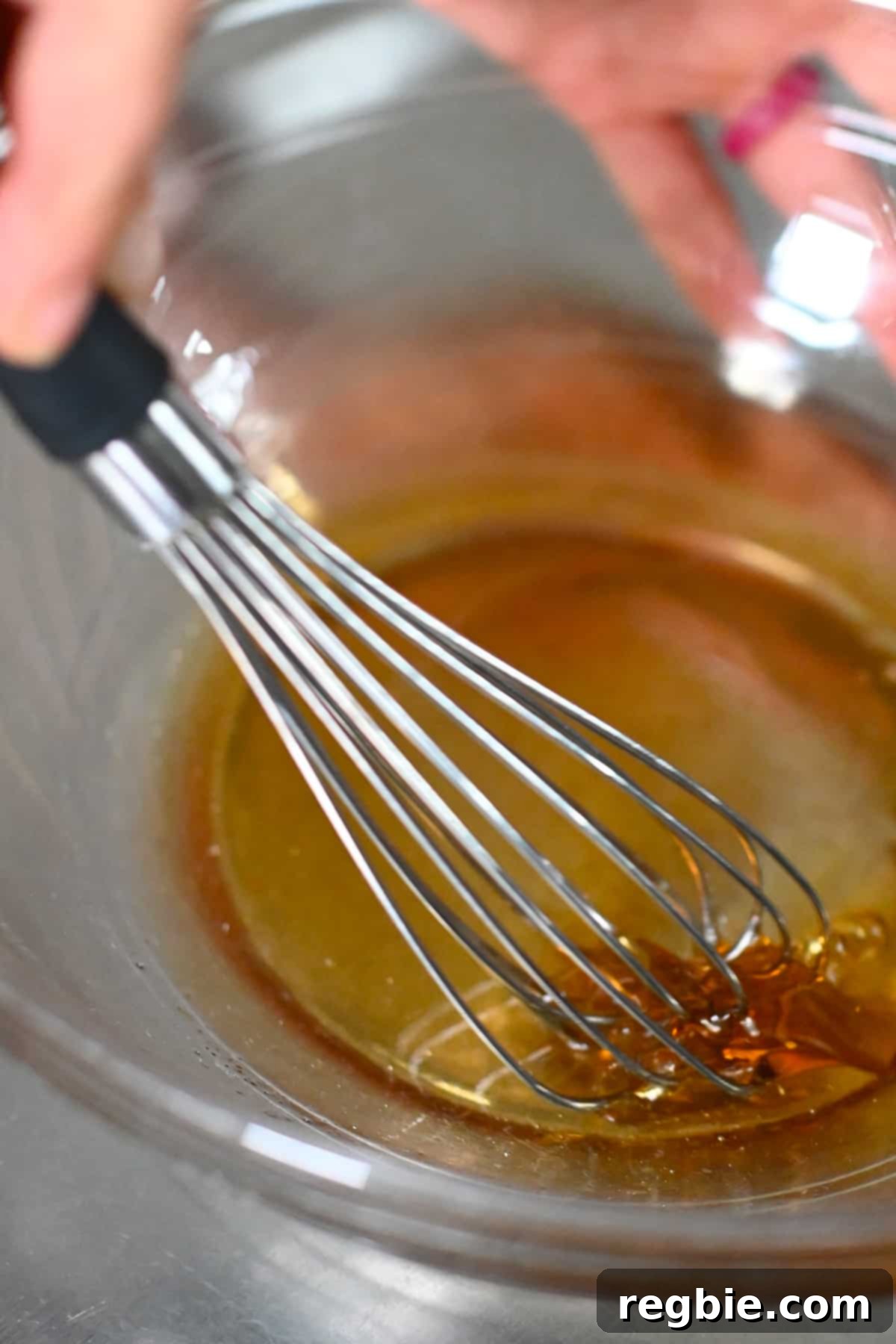Closeup of a whisk mixing melted coconut oil and maple syrup in a clear mixing bowl, forming the base for the pretzel crust.
