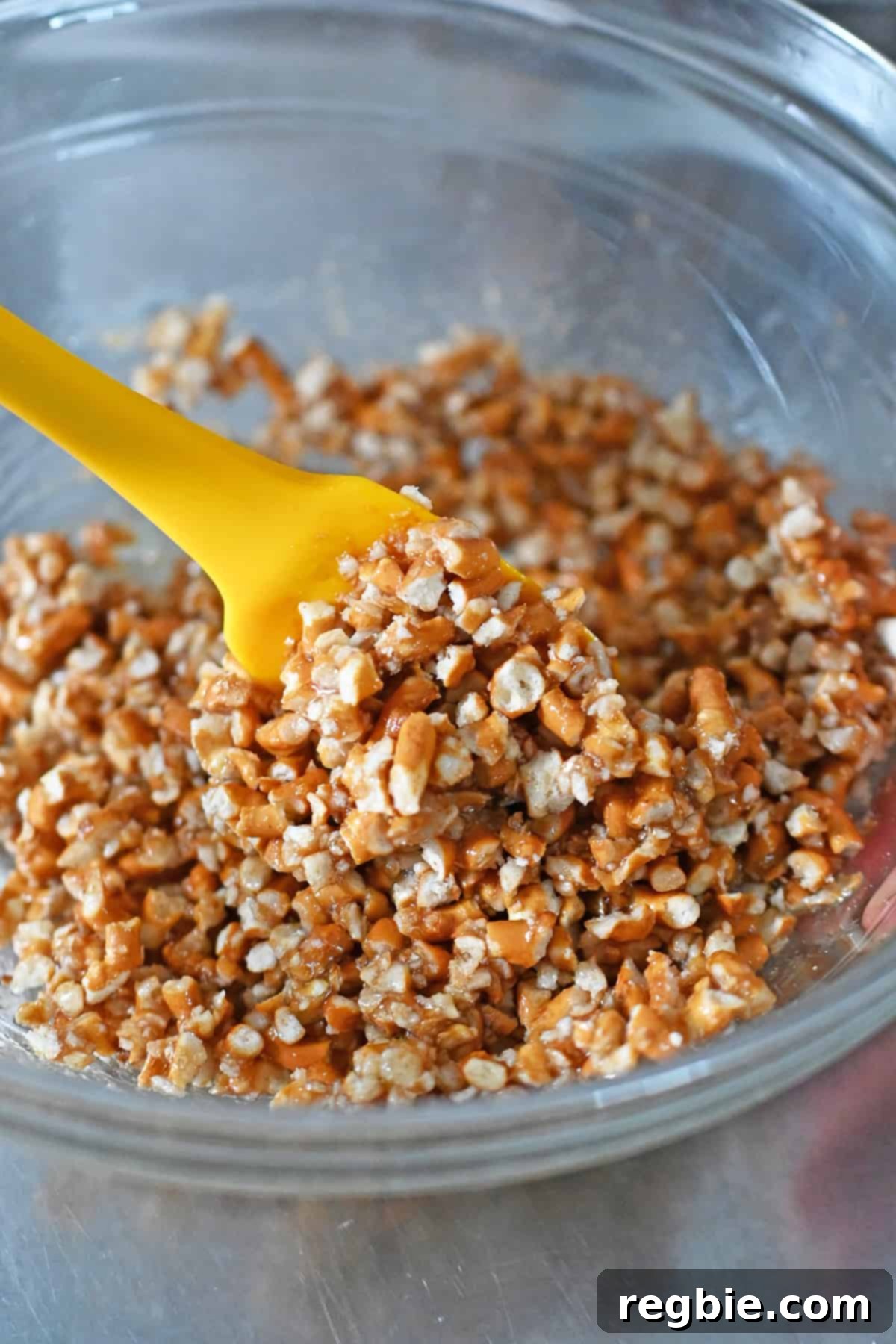 A yellow silicone spatula is mixing the pretzel crust for a grain-free and gluten-free strawberry pretzel salad, ensuring even coating of all ingredients.
