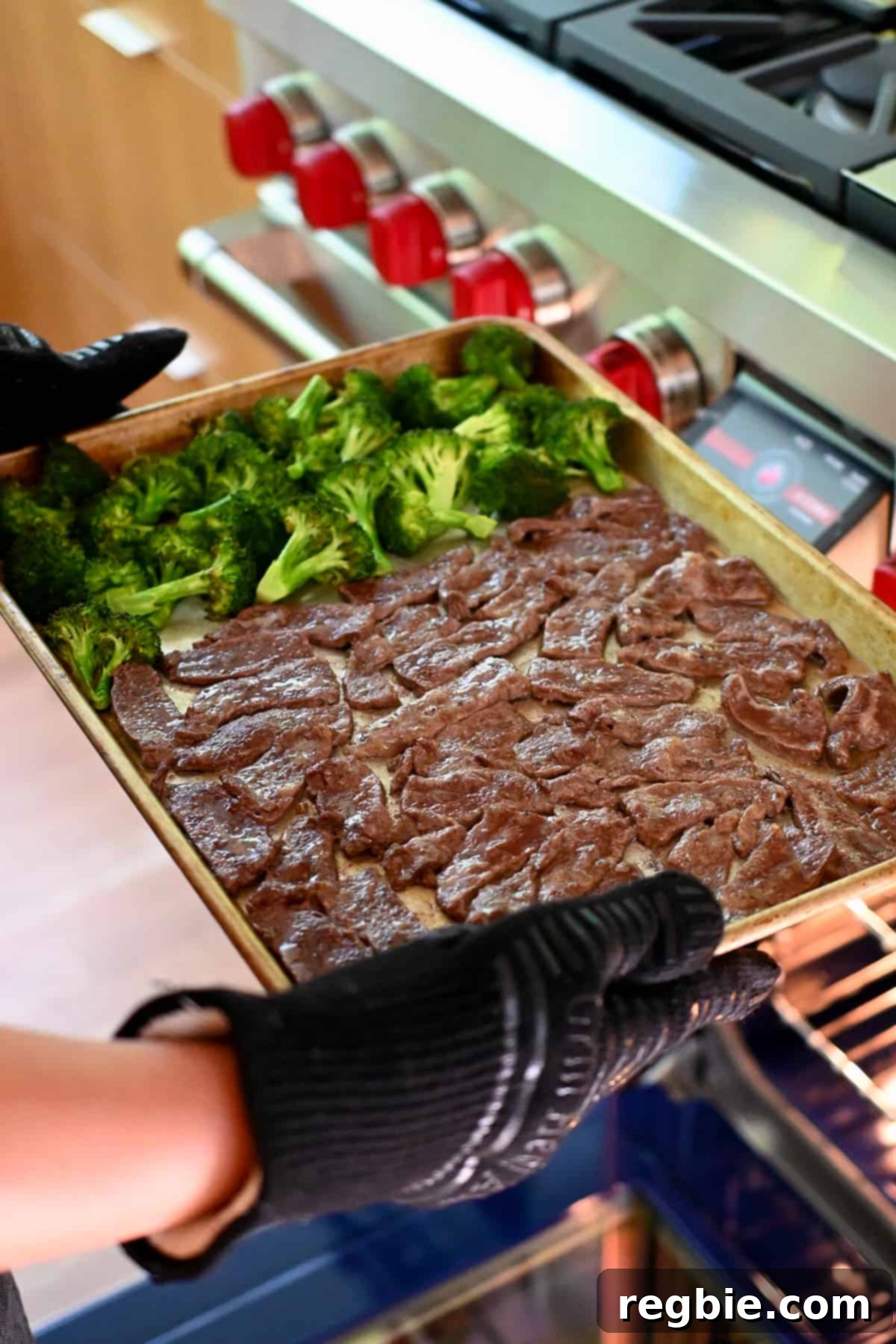 Someone is gently lifting a sheet pan, filled with partially cooked sliced beef and broccoli, out of the oven, ready for the sauce to be added.