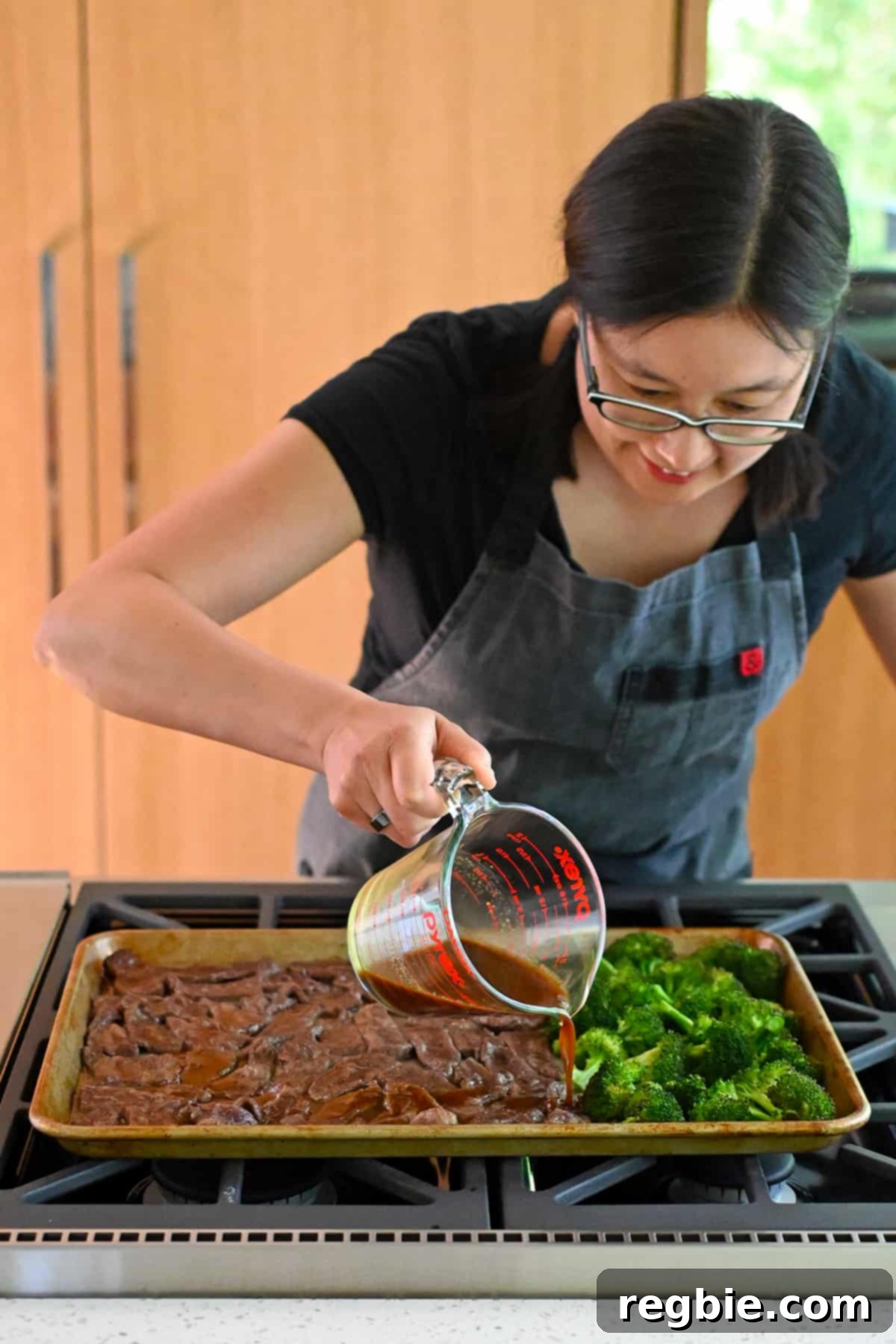 An Asian woman is carefully pouring a rich brown sauce evenly over the perfectly cooked sliced beef and broccoli on the sheet pan.