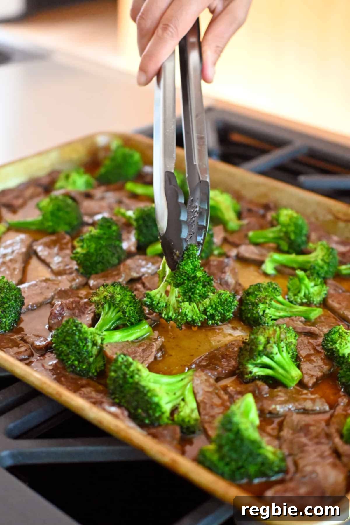 A pair of tongs is gently rearranging the sauced beef and broccoli on the sheet pan, ensuring a single layer before it goes under the broiler for finishing.