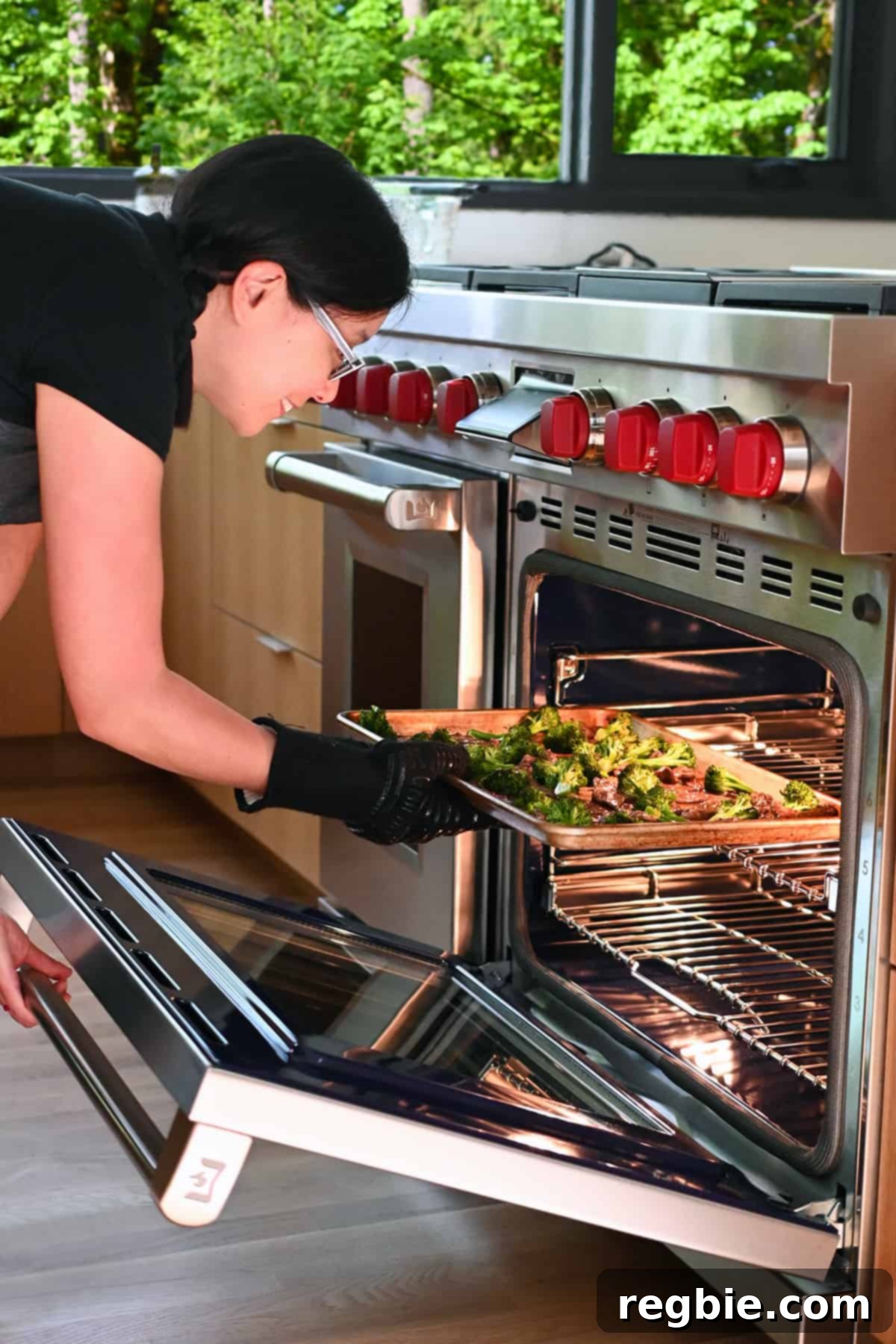 An Asian woman is carefully sliding the sheet pan, filled with beef and broccoli, under the broiler in an open oven to achieve a perfect finish.