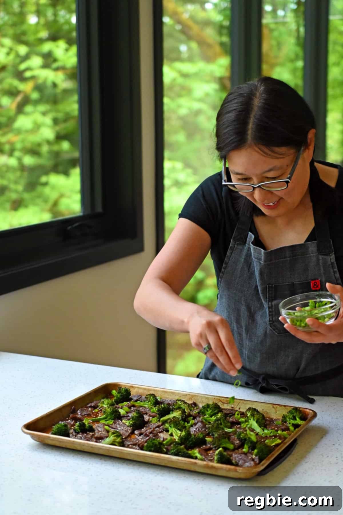 An Asian woman is gracefully sprinkling thinly sliced green onions over a rimmed baking sheet brimming with hot, delicious Chinese beef and broccoli.