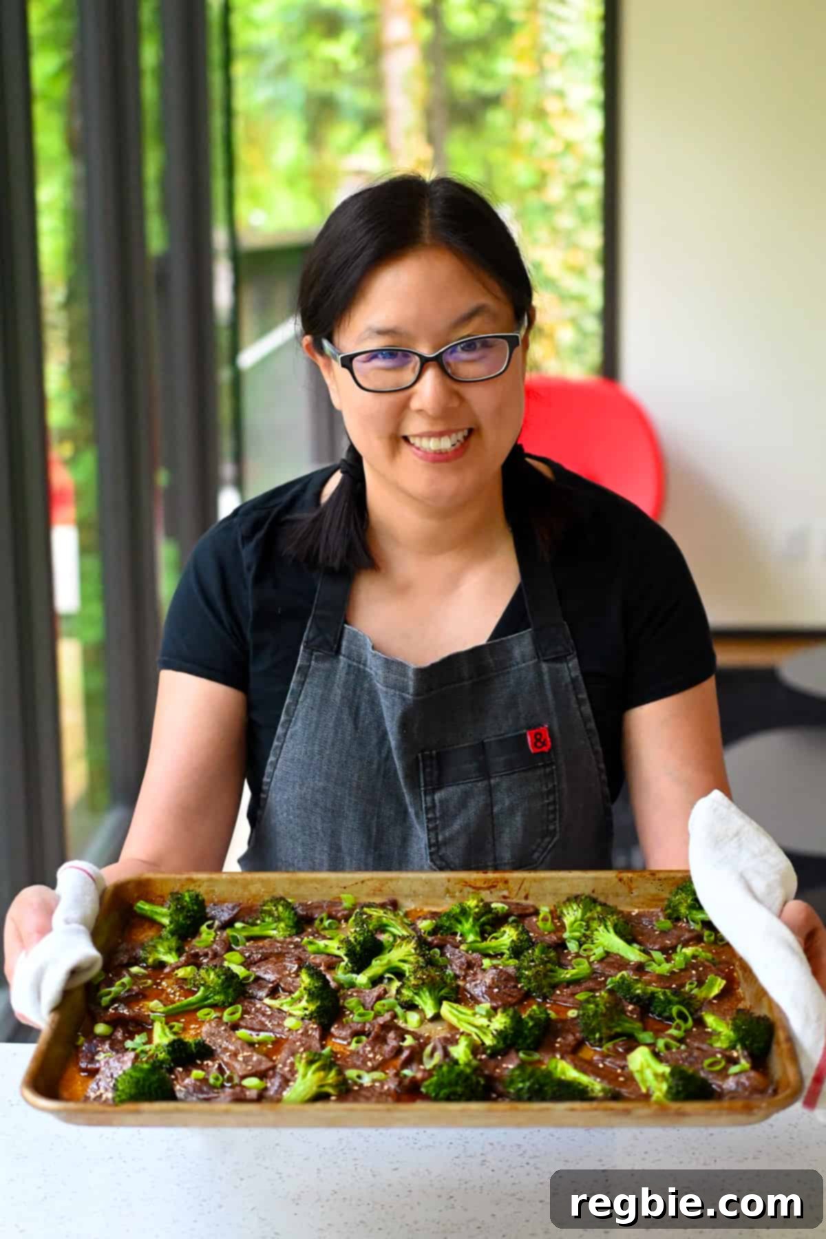 A smiling Asian woman happily holding a sheet pan filled with steaming Chinese beef and broccoli, fresh out of the oven, showcasing the dish's appeal.