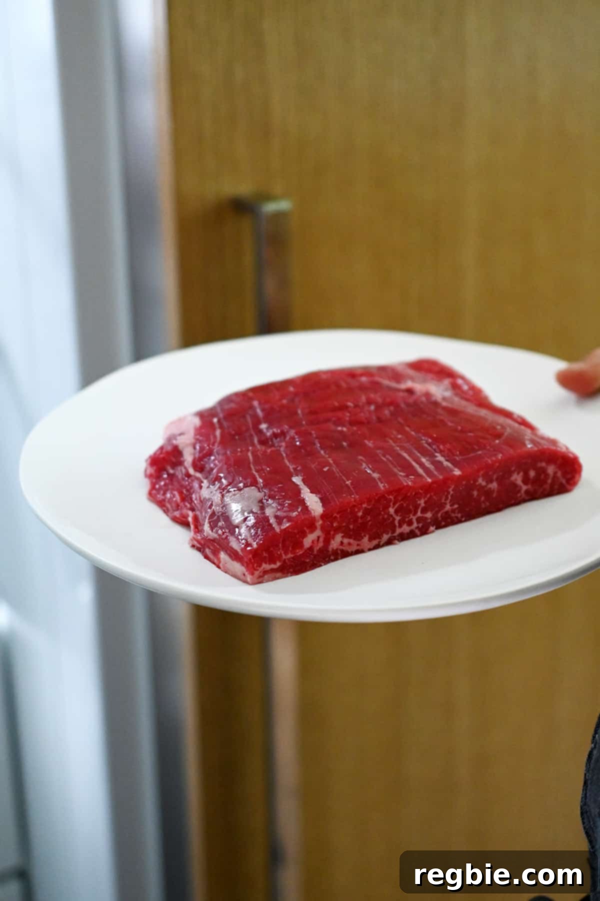 A hand is carefully placing a white plate with a raw piece of flank steak into the freezer, a key step for easier slicing.