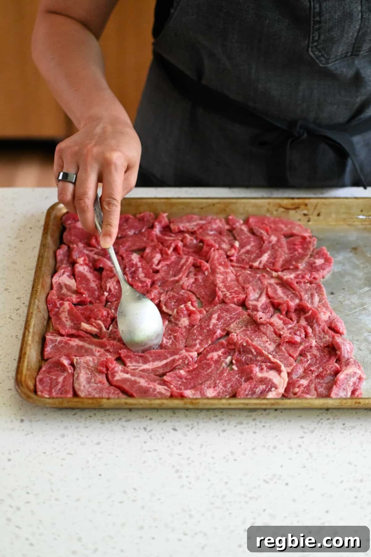 A spoon is meticulously arranging thinly sliced raw flank steak in a single layer on one half of a sheet pan, ensuring even cooking.