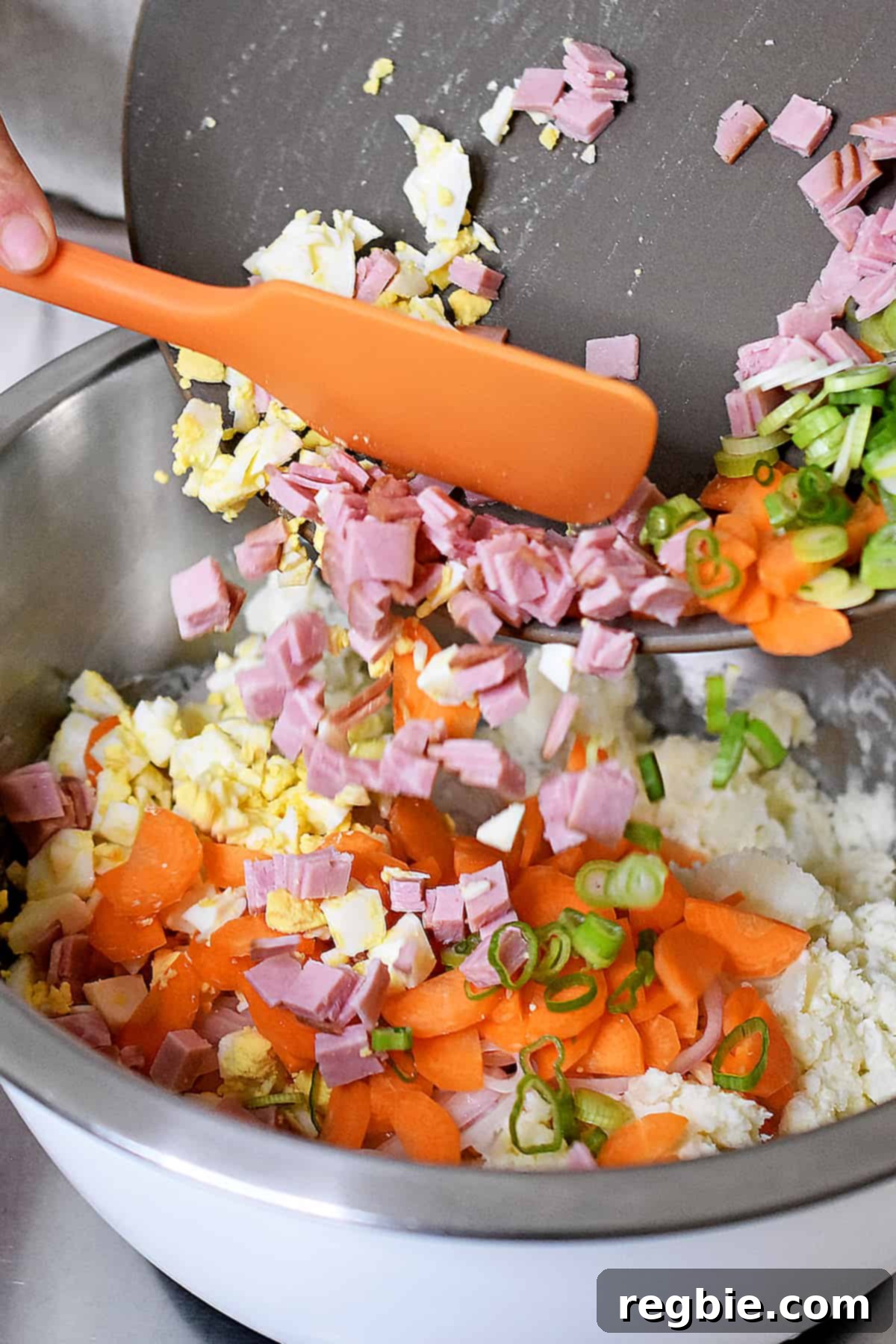 Adding diced ham, sliced carrots, hard boiled eggs, and sliced green onions to a bowl filled with mashed potatoes and dressing to complete the Japanese potato salad.
