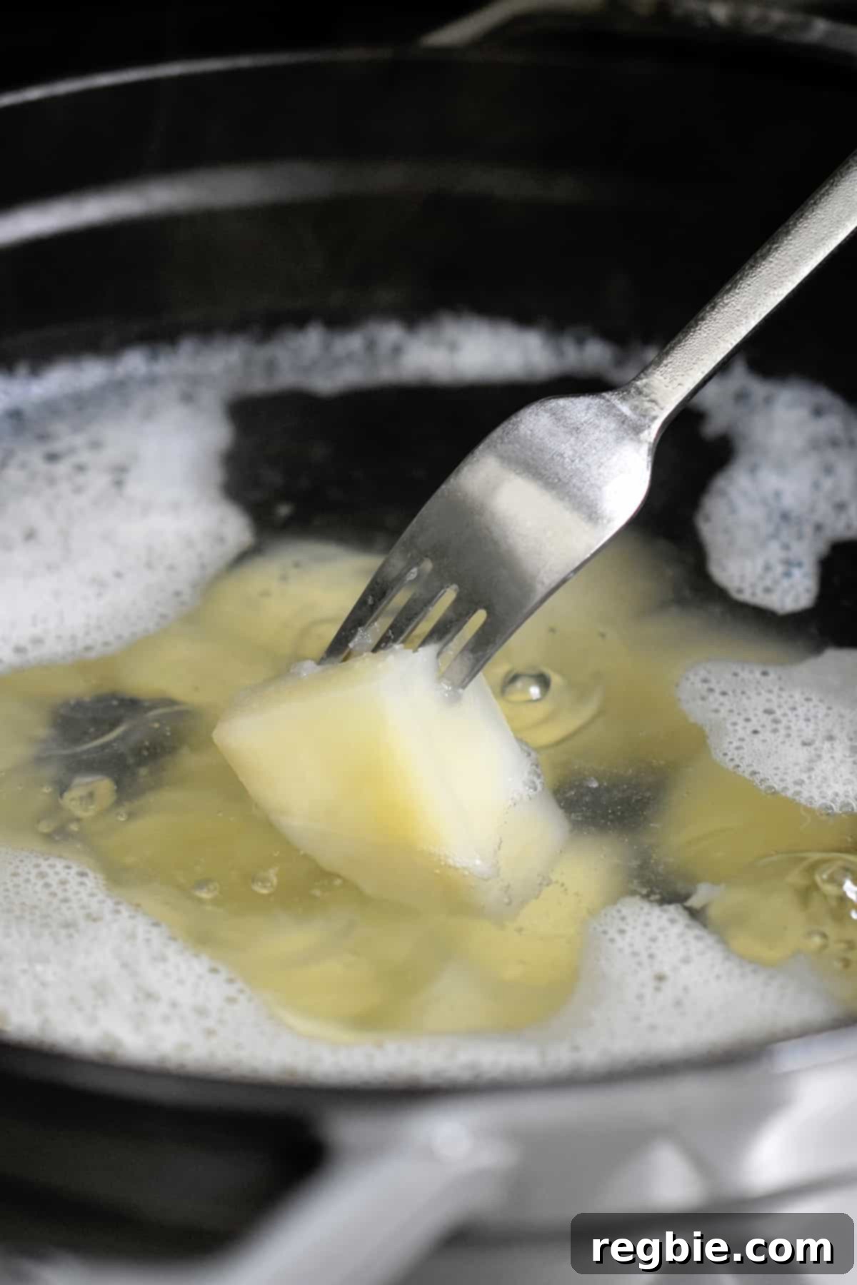 A fork is piercing a potato cube in a pot of boiling water, demonstrating its tenderness.