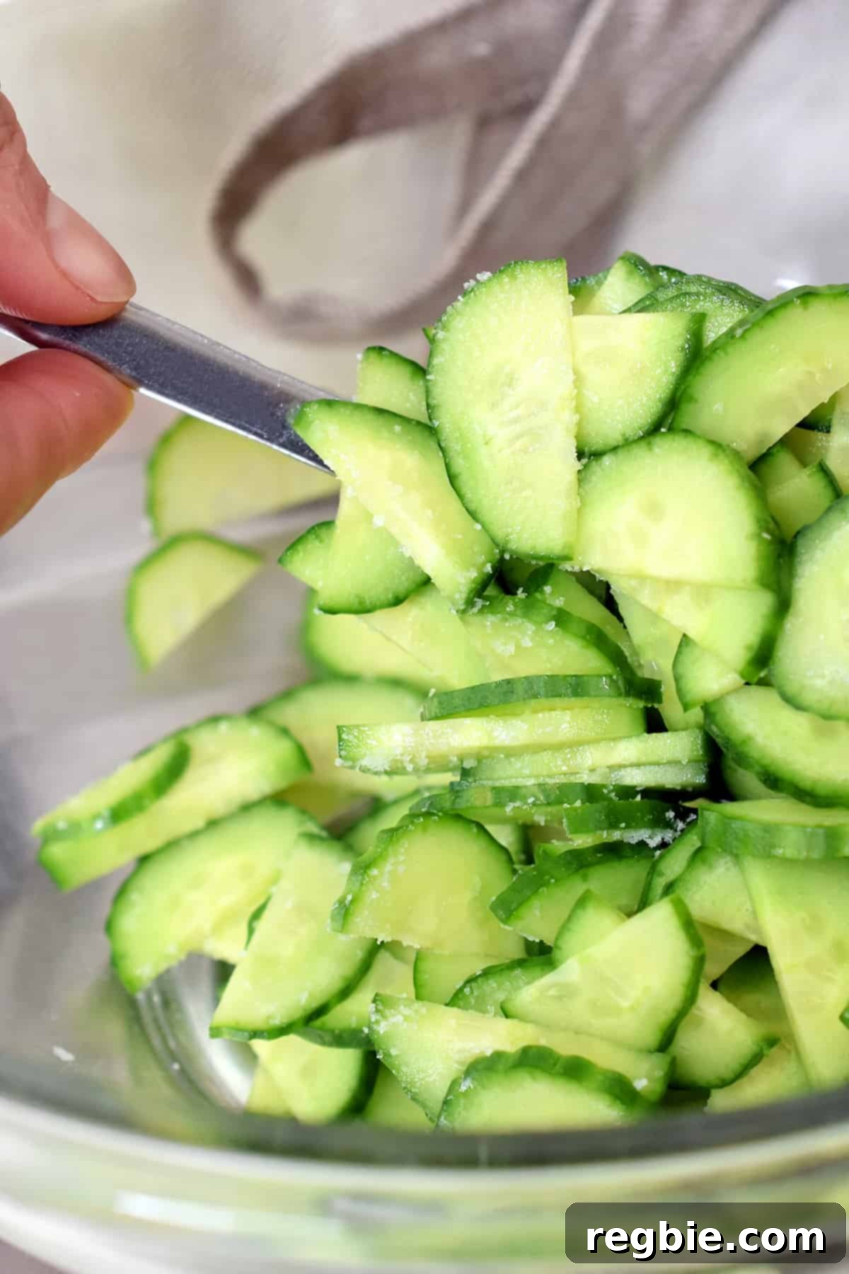 A closeup of thinly sliced Persian cucumbers with salt sprinkled on top in a glass bowl, starting the moisture-release process.