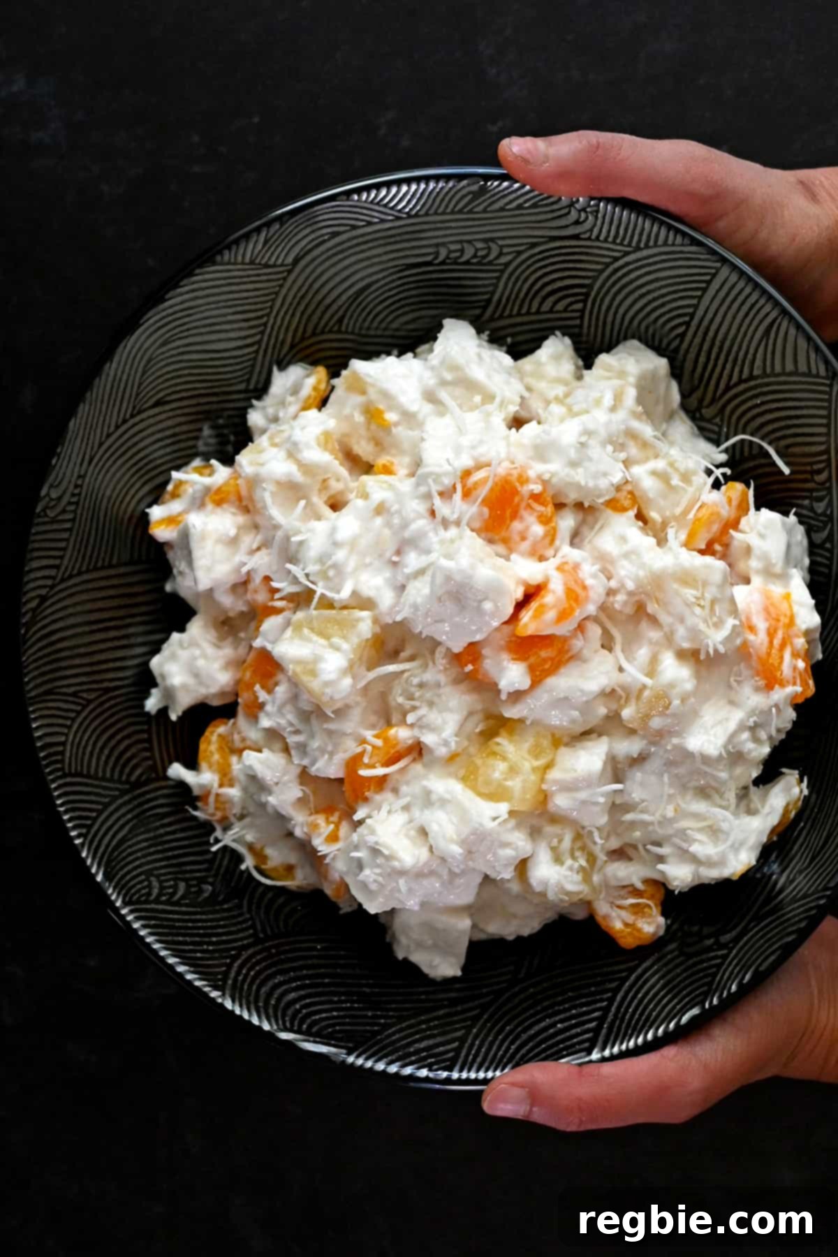 An overhead shot of two hands gently holding a sleek black serving bowl brimming with freshly prepared paleo ambrosia salad, ready to be presented.