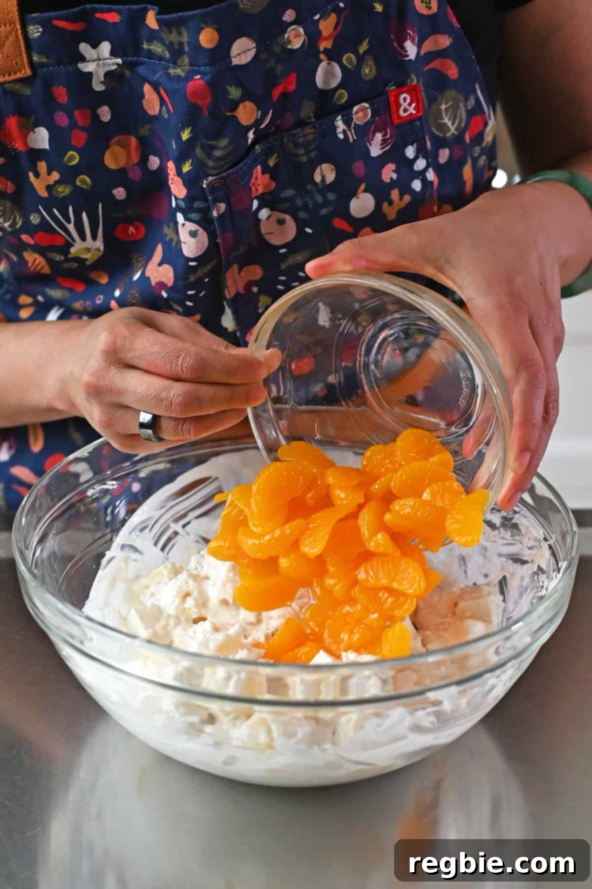 A visual of adding a bowl of perfectly drained canned mandarin oranges into the already mixed paleo ambrosia salad.