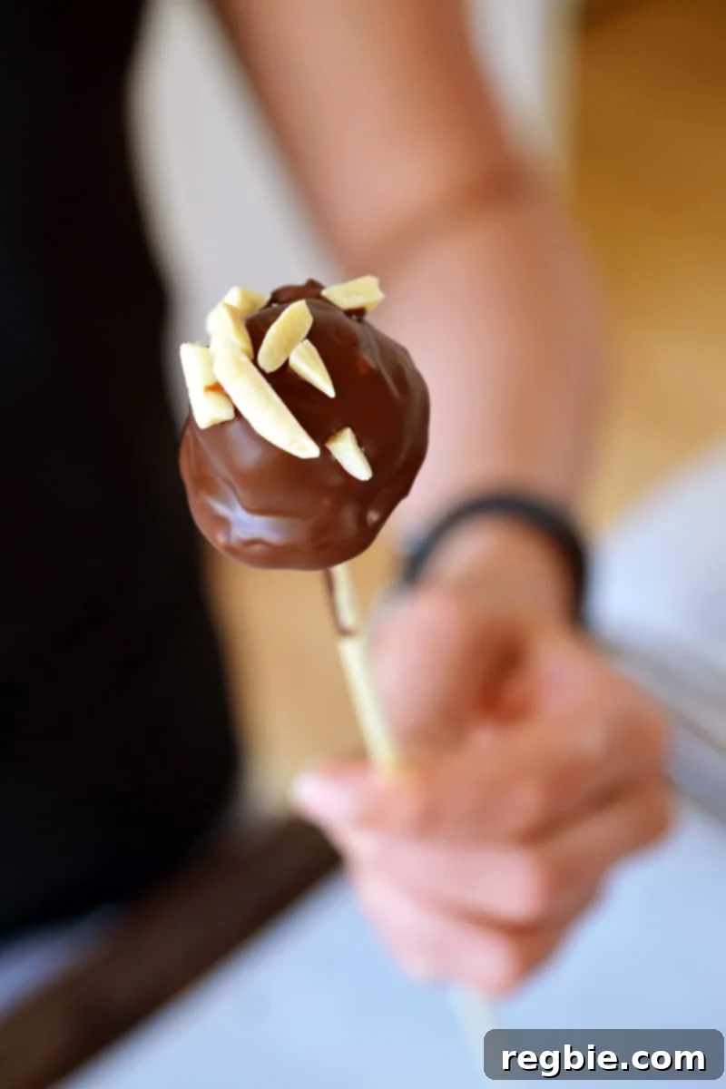 Guilt-Free Chunky Monkey Nice Cream Bonbons 17 A person in a stylish black shirt holds a delicious Chunky Monkey Ice Cream Bon Bon, perfectly coated in chocolate and sprinkled with nuts, on a slender wooden skewer.