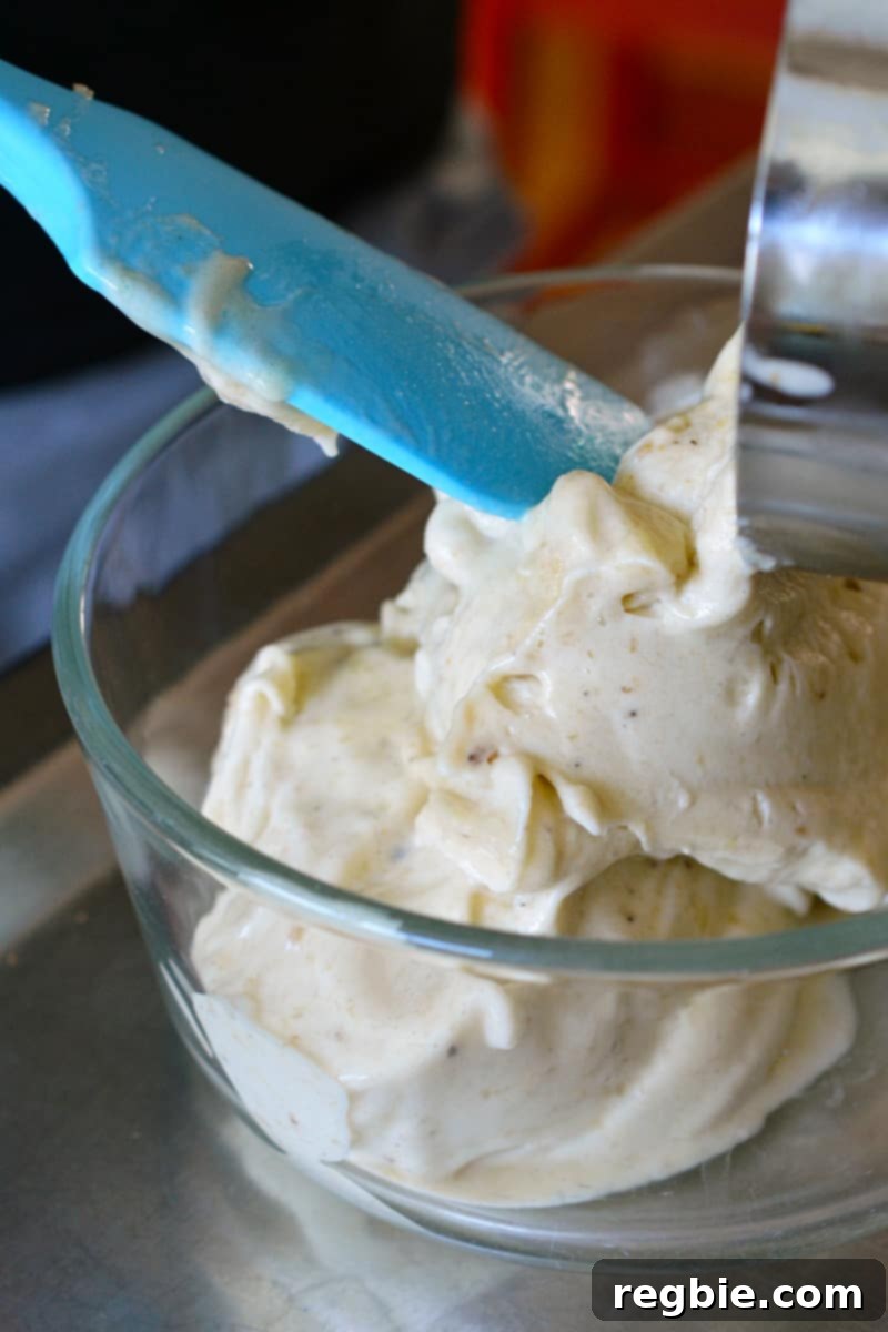 Guilt-Free Chunky Monkey Nice Cream Bonbons 7 A smooth, light-colored vegan banana nice cream is being carefully transferred into a clear glass container with a cheerful blue silicone spatula, preparing for its chilling phase.