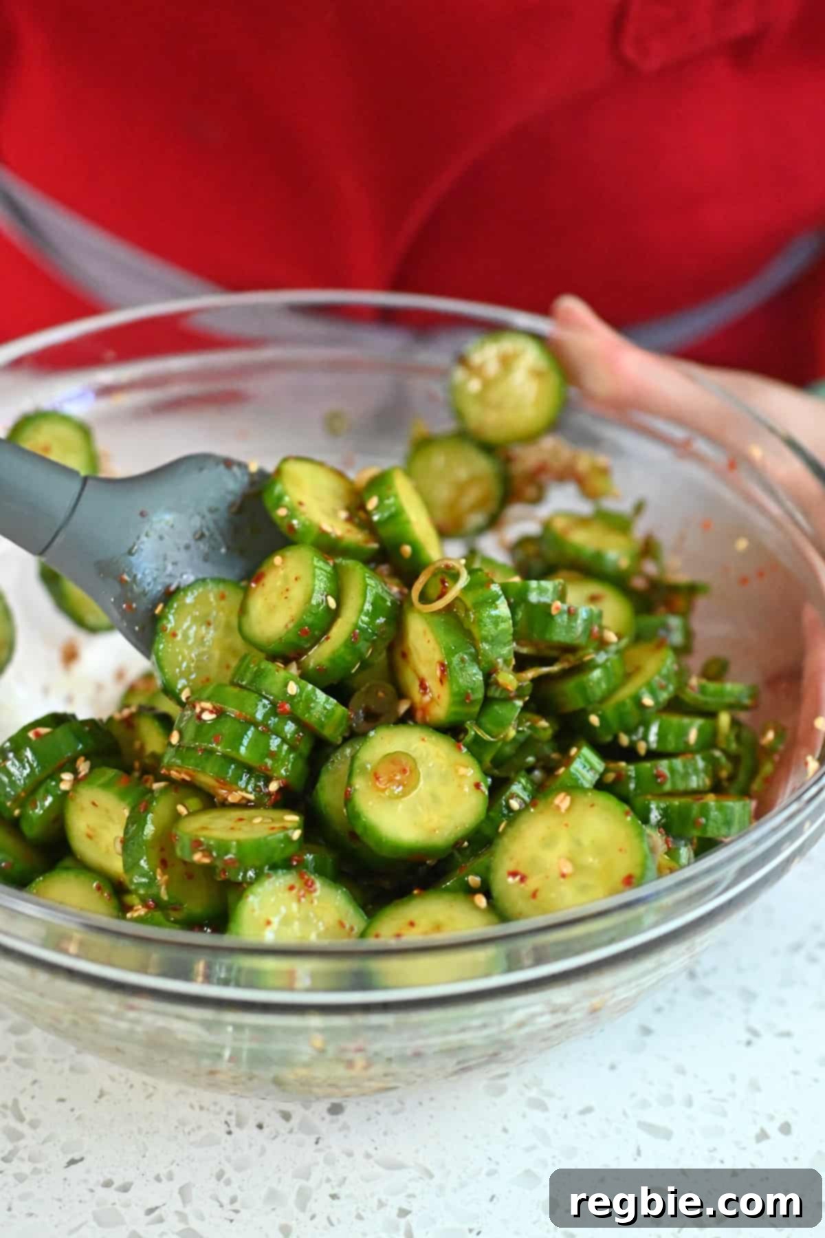 A close-up shot of Korean cucumber salad being tossed with a gray silicone spatula in a clear glass bowl, ensuring all ingredients are evenly mixed.