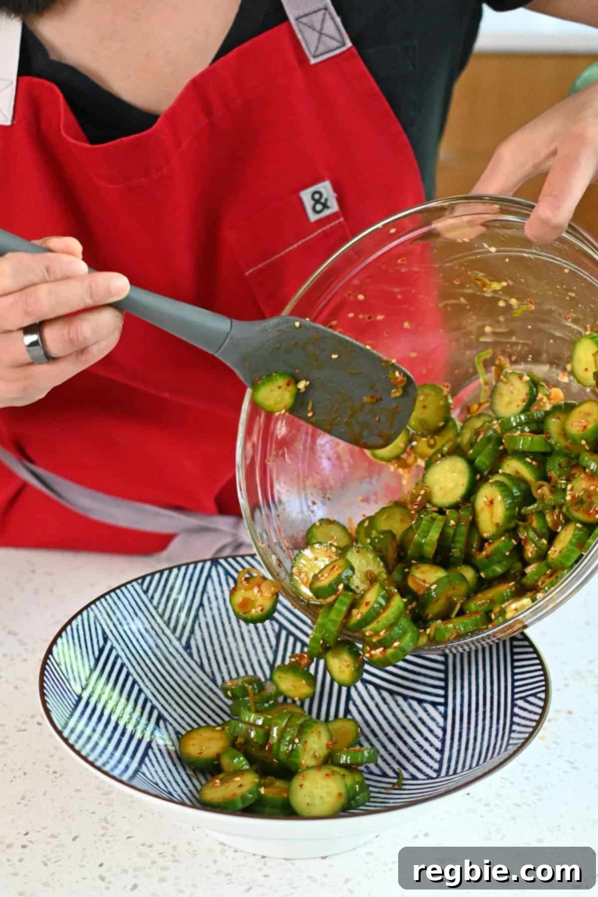 A cook expertly transferring freshly prepared Korean cucumber salad from a clear glass mixing bowl to an elegant blue and white serving platter.