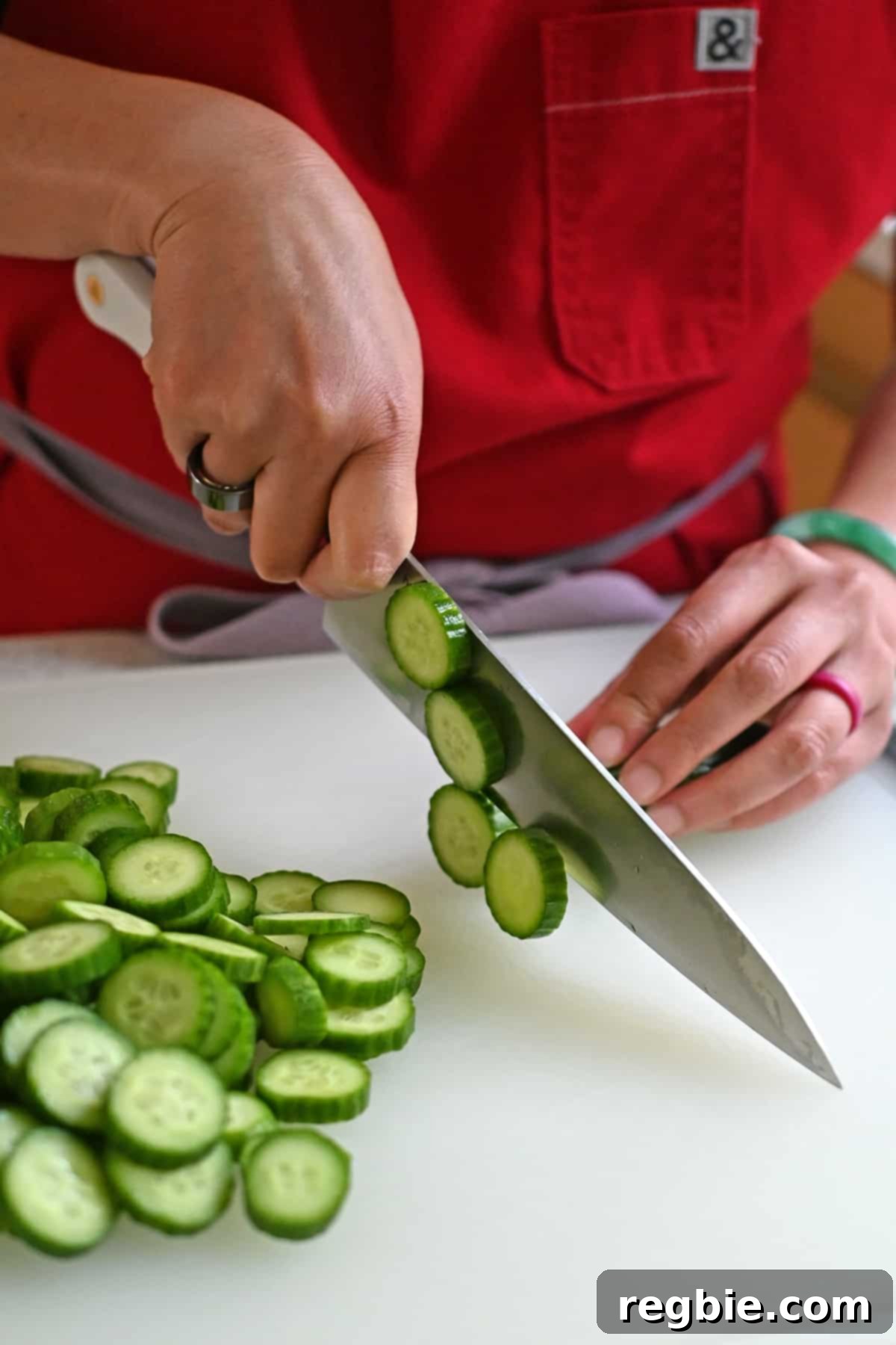 A person in a red apron meticulously cutting fresh Persian cucumbers into thin, uniform slices using a professional chef's knife on a cutting board.