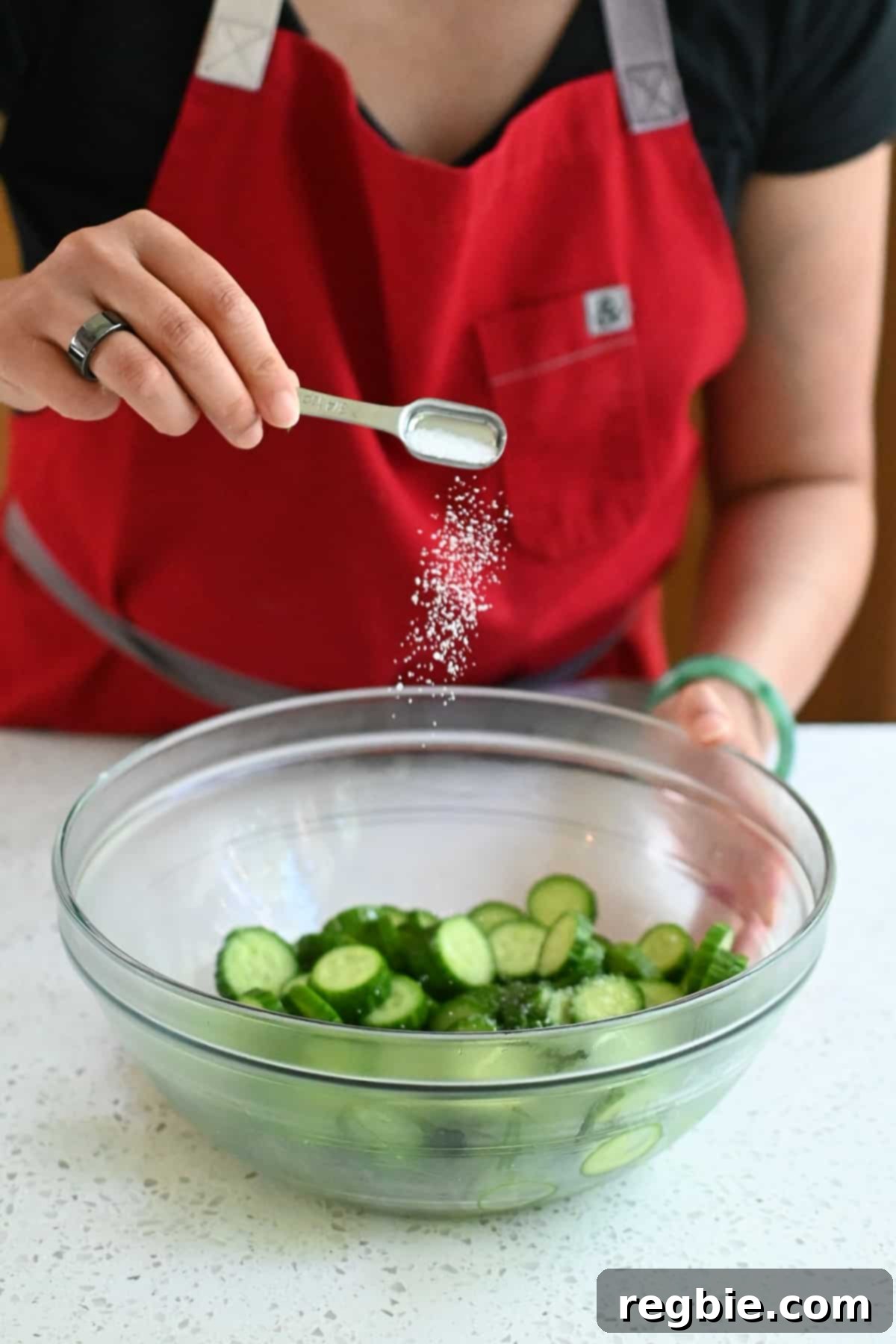 A hand adding a sprinkle of Diamond Crystal kosher salt to a bowl filled with freshly sliced Persian cucumbers, preparing them for the Korean cucumber salad.