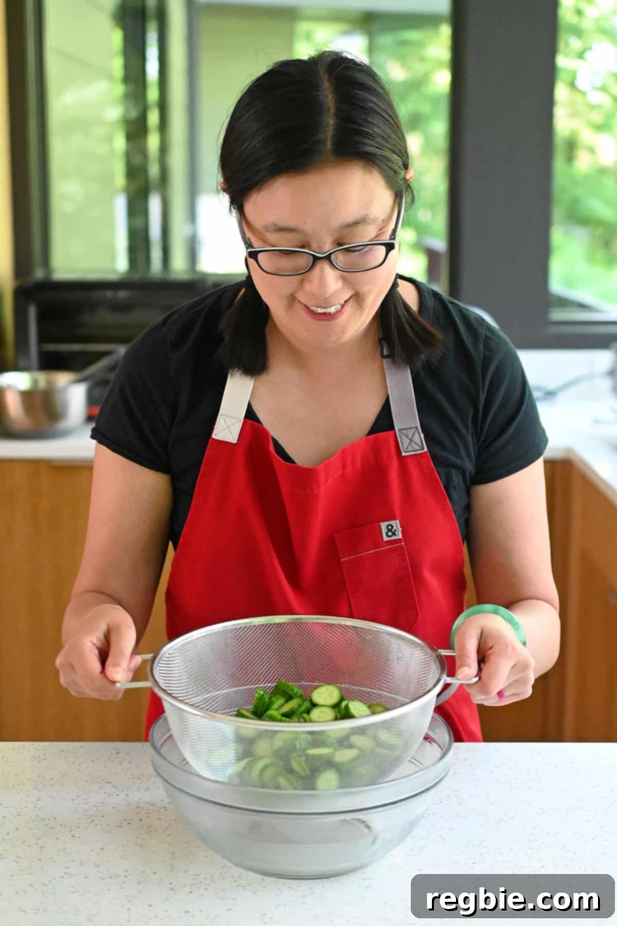 A colander brimming with salted, sliced cucumbers being carefully placed into a larger glass mixing bowl, ready for the draining process.