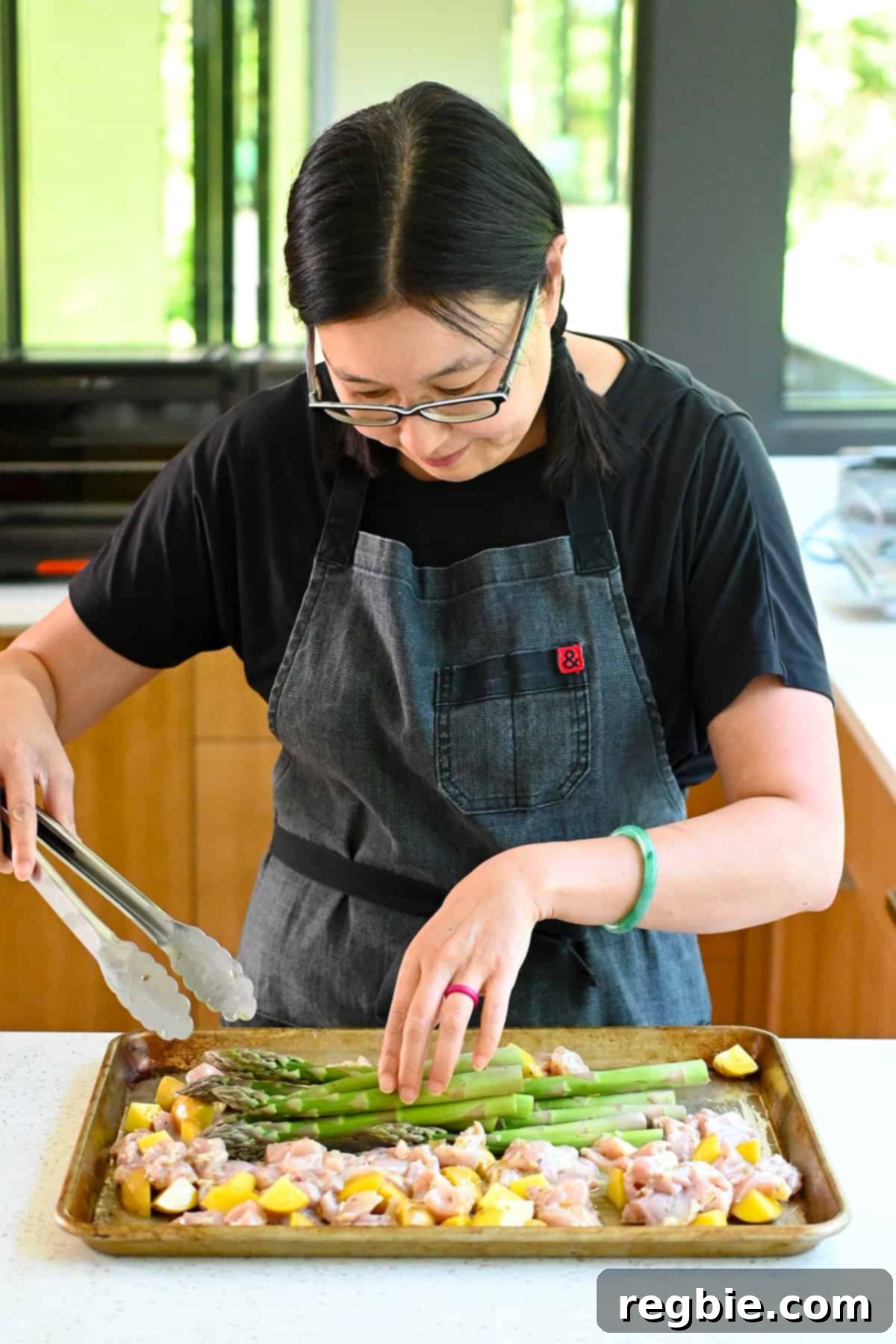 Zesty Mediterranean Sheet Pan Chicken 11 An Asian woman gently places fresh asparagus spears onto the sheet pan, alongside the marinated chicken and potatoes, ready for a thorough coating in the flavorful Greek dressing.