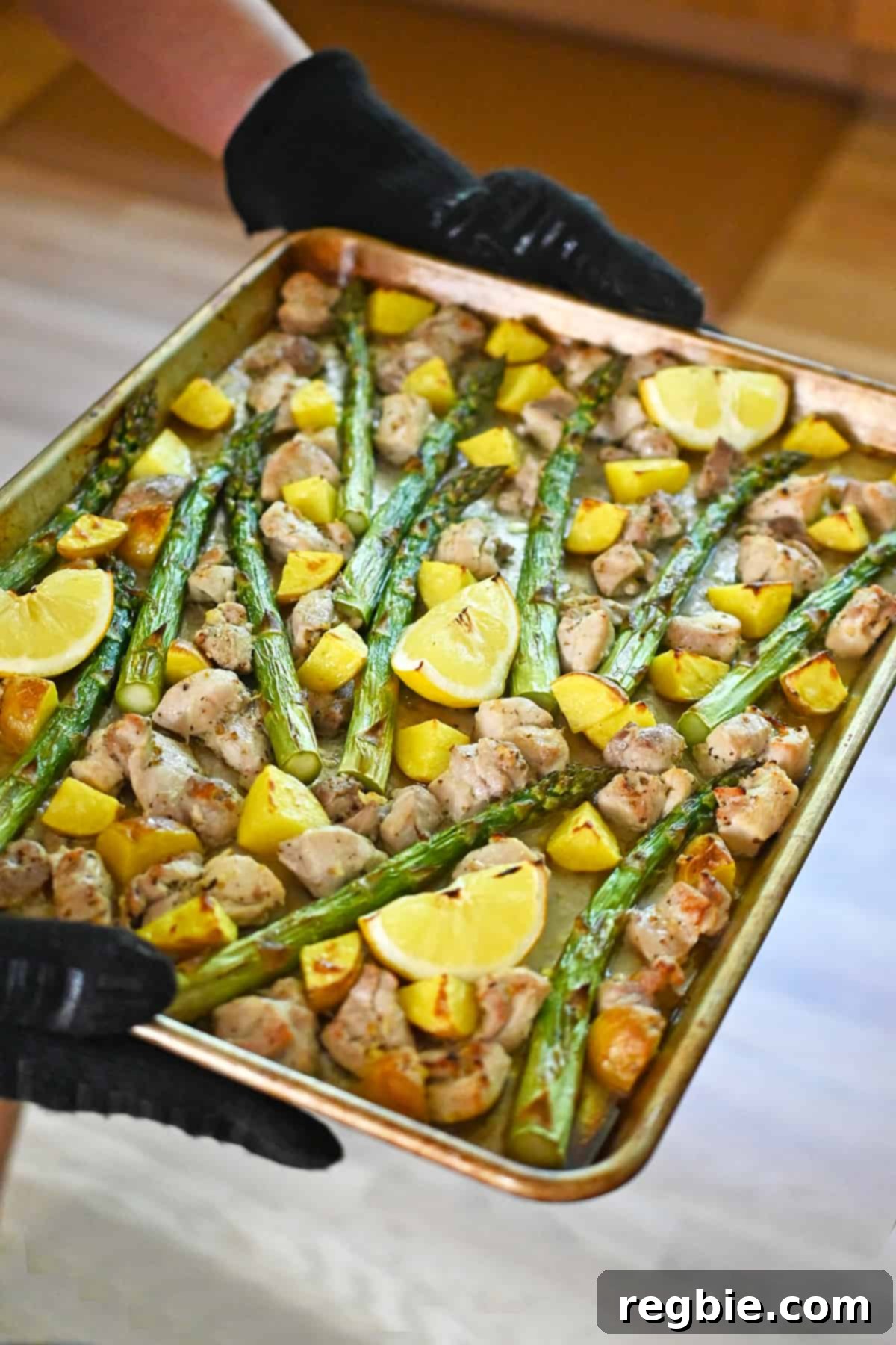 Zesty Mediterranean Sheet Pan Chicken 14 Hands, protected by oven mitts, retrieve a sheet pan brimming with beautifully golden-brown chicken, roasted potatoes, tender asparagus, and perfectly charred lemon wedges from the oven, signifying completion.