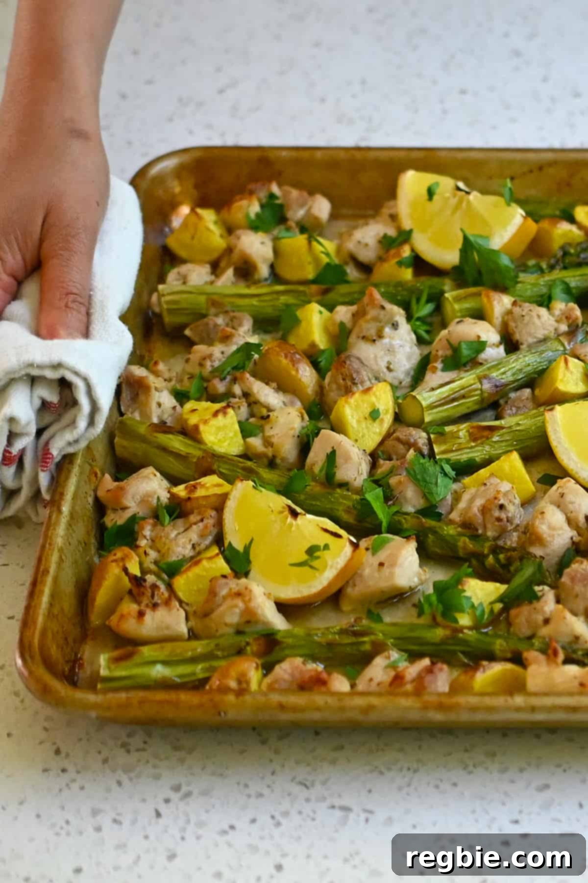 Zesty Mediterranean Sheet Pan Chicken 17 A warm, inviting hand holds a sheet pan laden with a delicious Greek chicken, asparagus, potatoes, and lemon wedges, ready to be served, complemented by a crisp white kitchen towel.