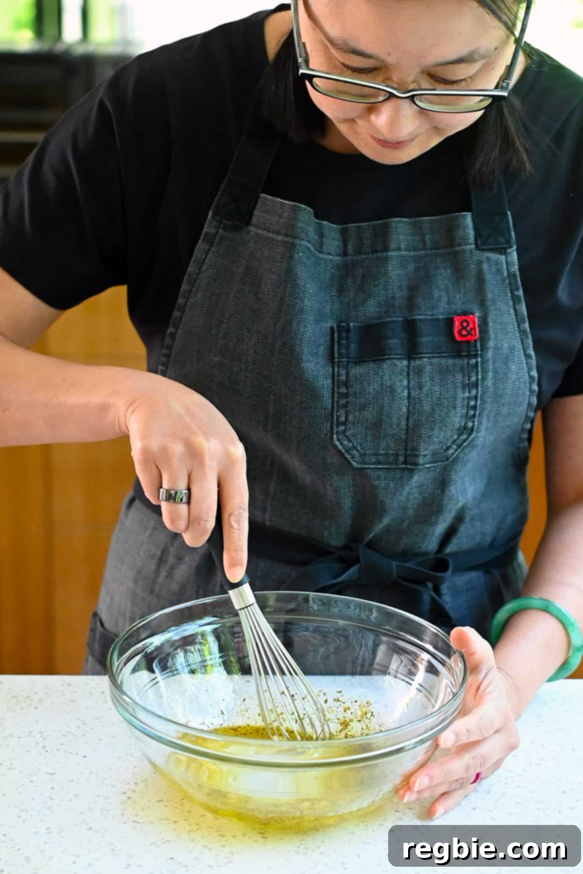 Zesty Mediterranean Sheet Pan Chicken 7 An Asian woman gracefully whisks the Greek-inspired marinade in a clear glass mixing bowl, creating a perfectly blended and aromatic dressing.