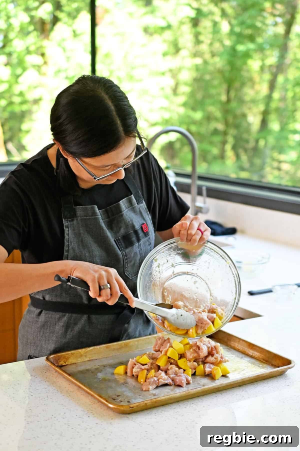 Zesty Mediterranean Sheet Pan Chicken 10 An Asian woman carefully transfers the marinated raw chicken and potatoes from the mixing bowl onto a greased rimmed baking sheet, ensuring an even spread for optimal cooking.
