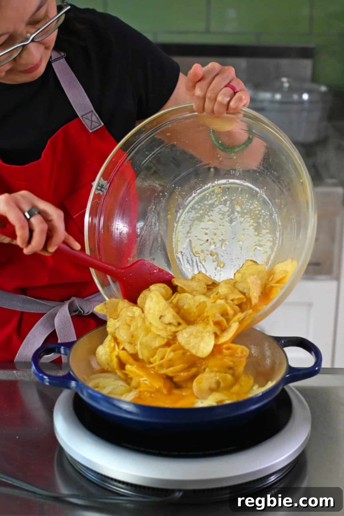Crispy Potato Chip Tortilla 11 An Asian woman in glasses is adding a bowl of whisked eggs and potato chips to a blue skillet with sautéed onions to make a quick Spanish tortilla.