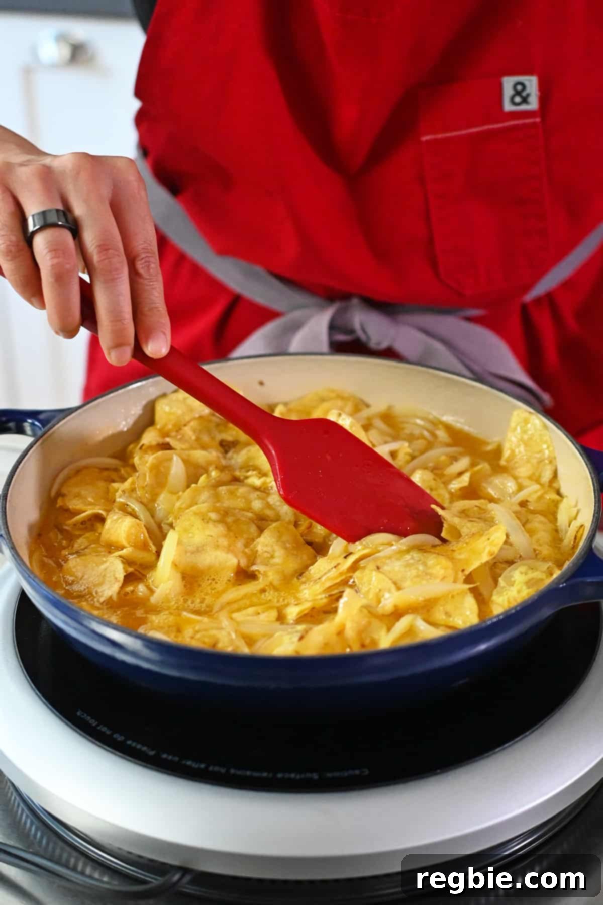 Crispy Potato Chip Tortilla 12 A red silicone spatula is rearranging potato chips and whisked raw eggs in a blue skillet so the top is a smooth layer.