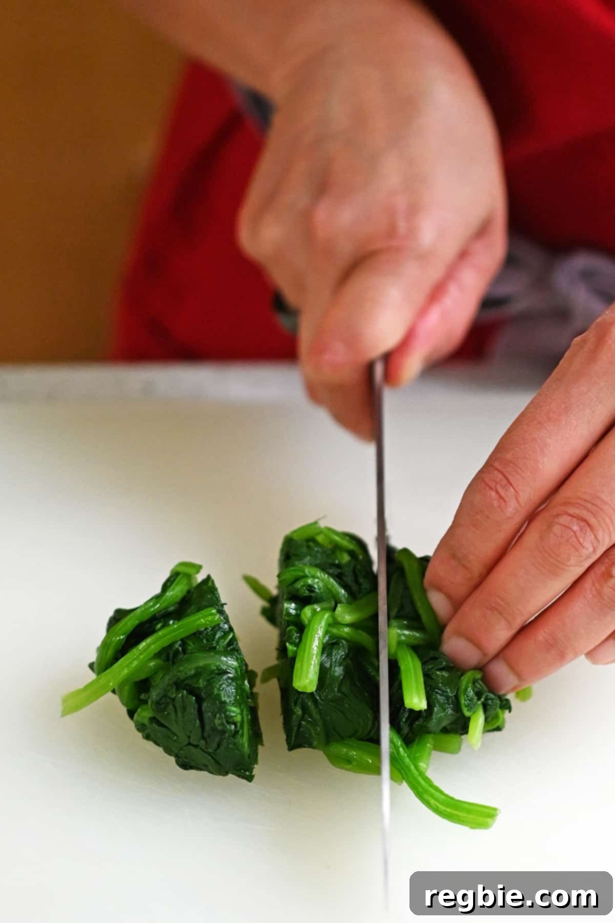 A chef's knife slices through a pile of cooked and thoroughly squeezed-dry spinach, dividing it into three manageable sections on a cutting board.