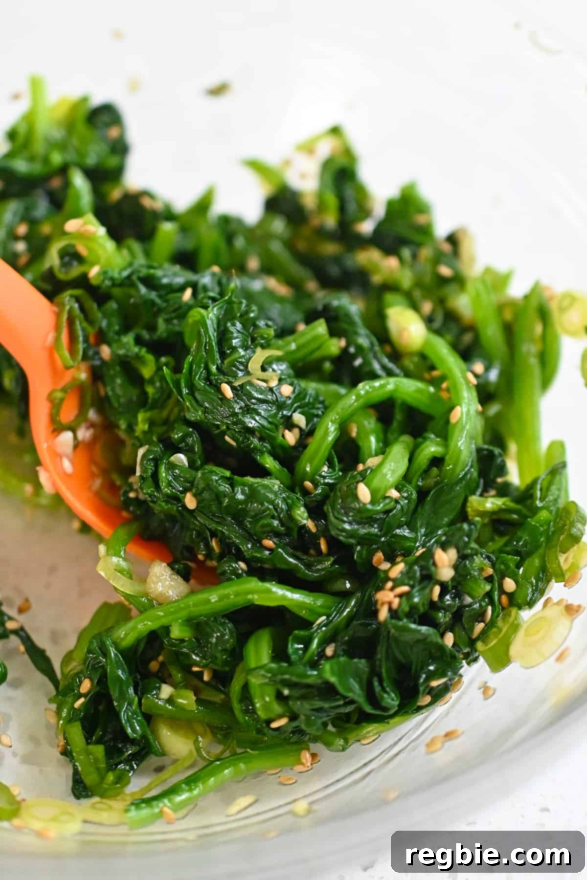 A close-up shot of a silicone spatula thoroughly mixing the Korean spinach side dish in a clear glass bowl, ensuring even distribution of flavors.