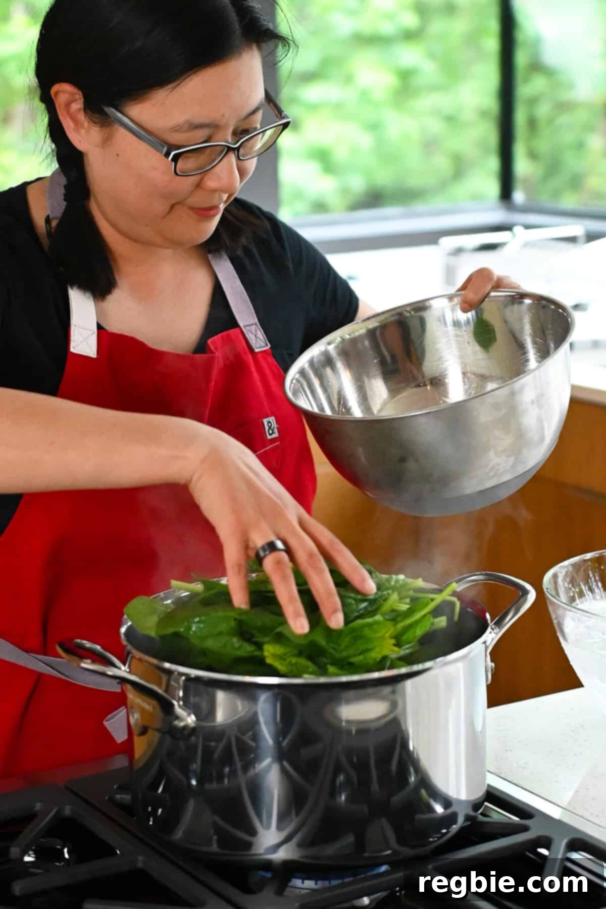 An Asian woman in a red apron gently adds fresh, raw spinach into a large pot of vigorously boiling water, preparing it for the Korean side dish.