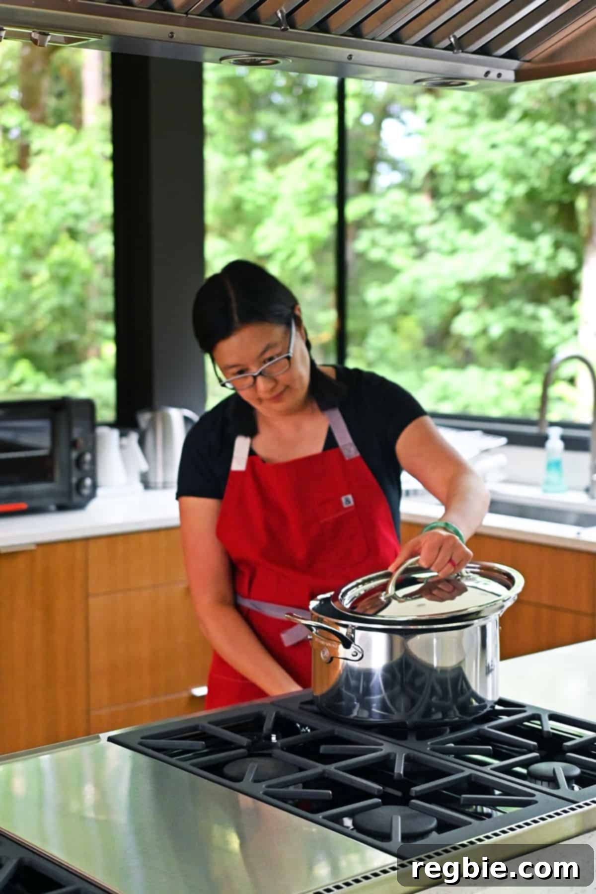 An Asian woman in a red apron gently places a lid on a large stockpot that is heating on a stovetop, preparing to blanch spinach.