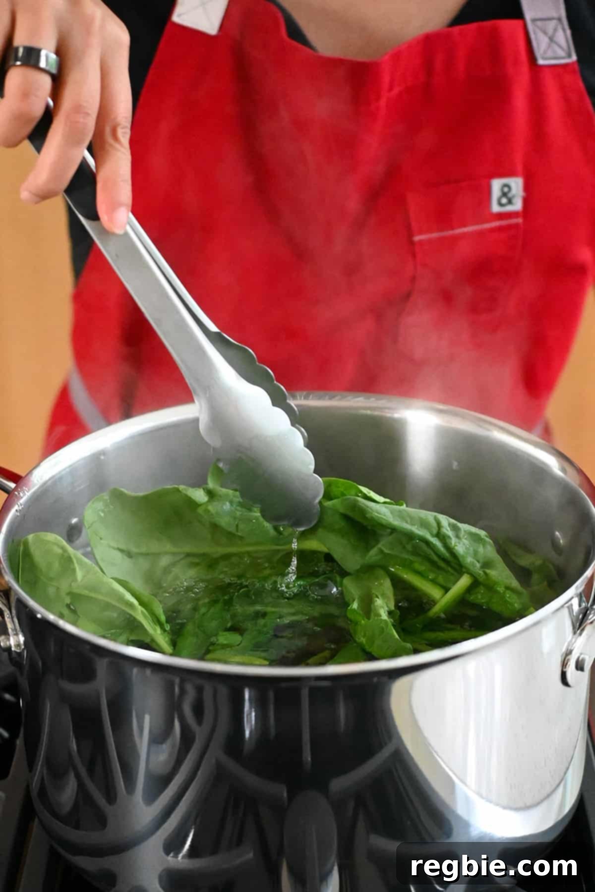 A pair of tongs carefully adds fresh, raw spinach into a large pot of boiling water, initiating the cooking process for a Korean spinach side dish.