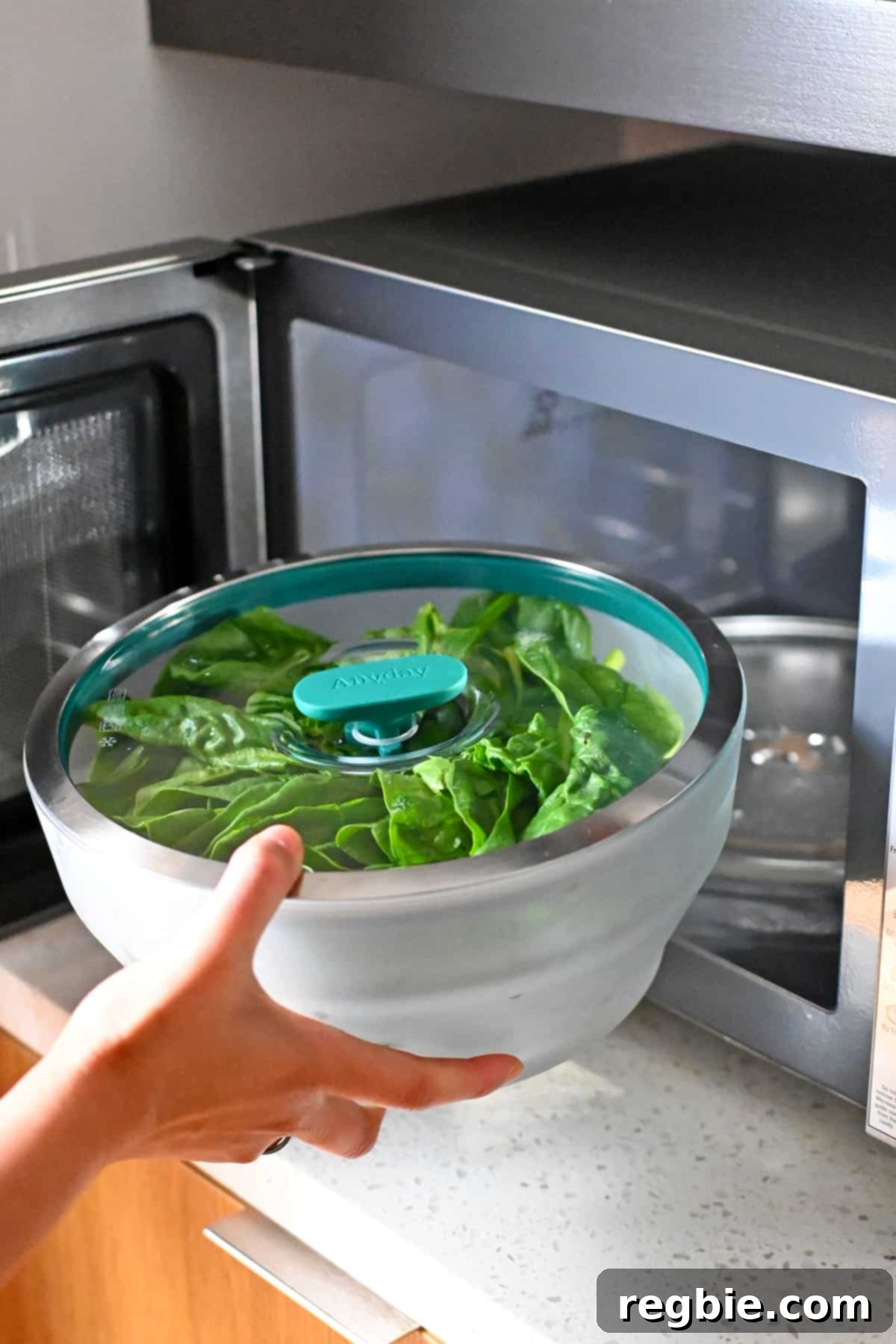 A hand gently places an extra-large, clear glass Anyday microwave container filled with fresh raw spinach into an open microwave oven.