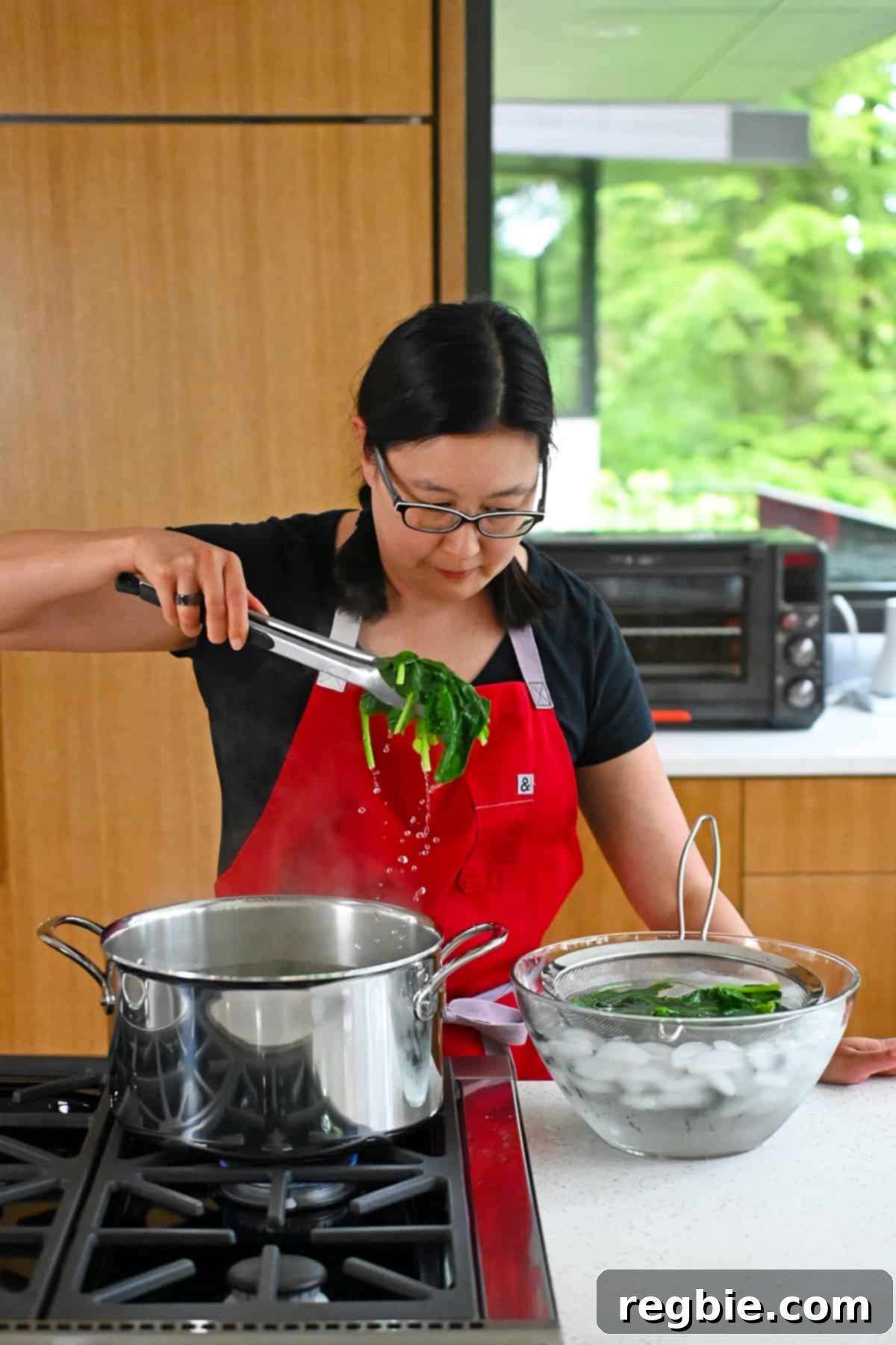 An Asian woman expertly transfers cooked spinach from a pot of boiling water directly into a glass bowl filled with ice water and a strainer, to cool it rapidly.