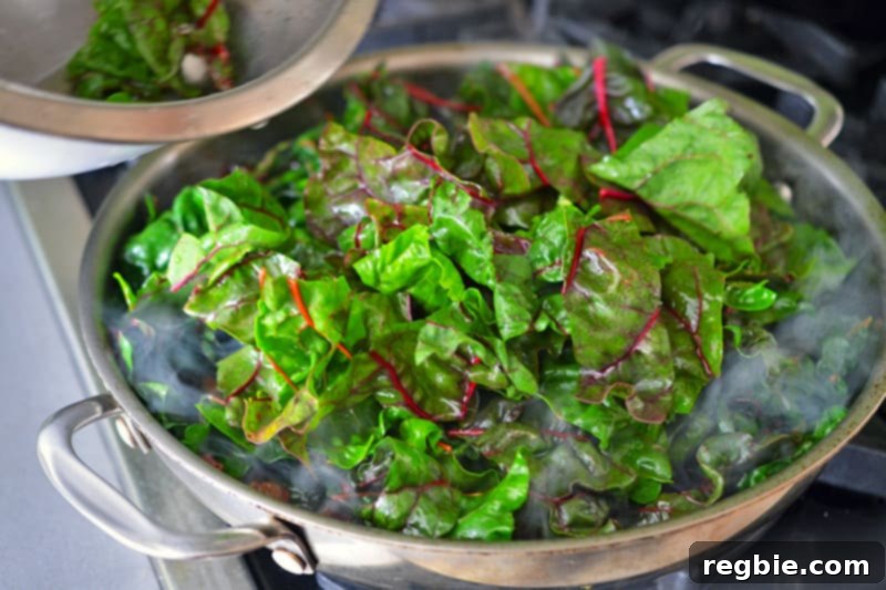 A large quantity of fresh Swiss chard leaves is being added in batches to a hot pan, beginning the wilting process as they touch the heat.