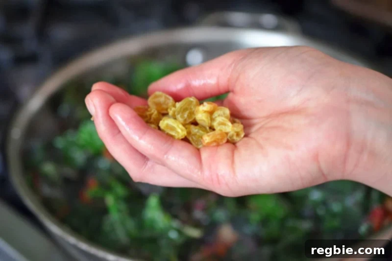 A hand holds a handful of plumped golden raisins, poised to add them to the pan with the sautéed Swiss chard and other flavorful ingredients.