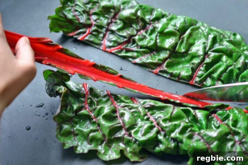A chef's knife is shown cutting out the thick stems from vibrant rainbow Swiss chard leaves on a wooden cutting board, separating the usable parts.